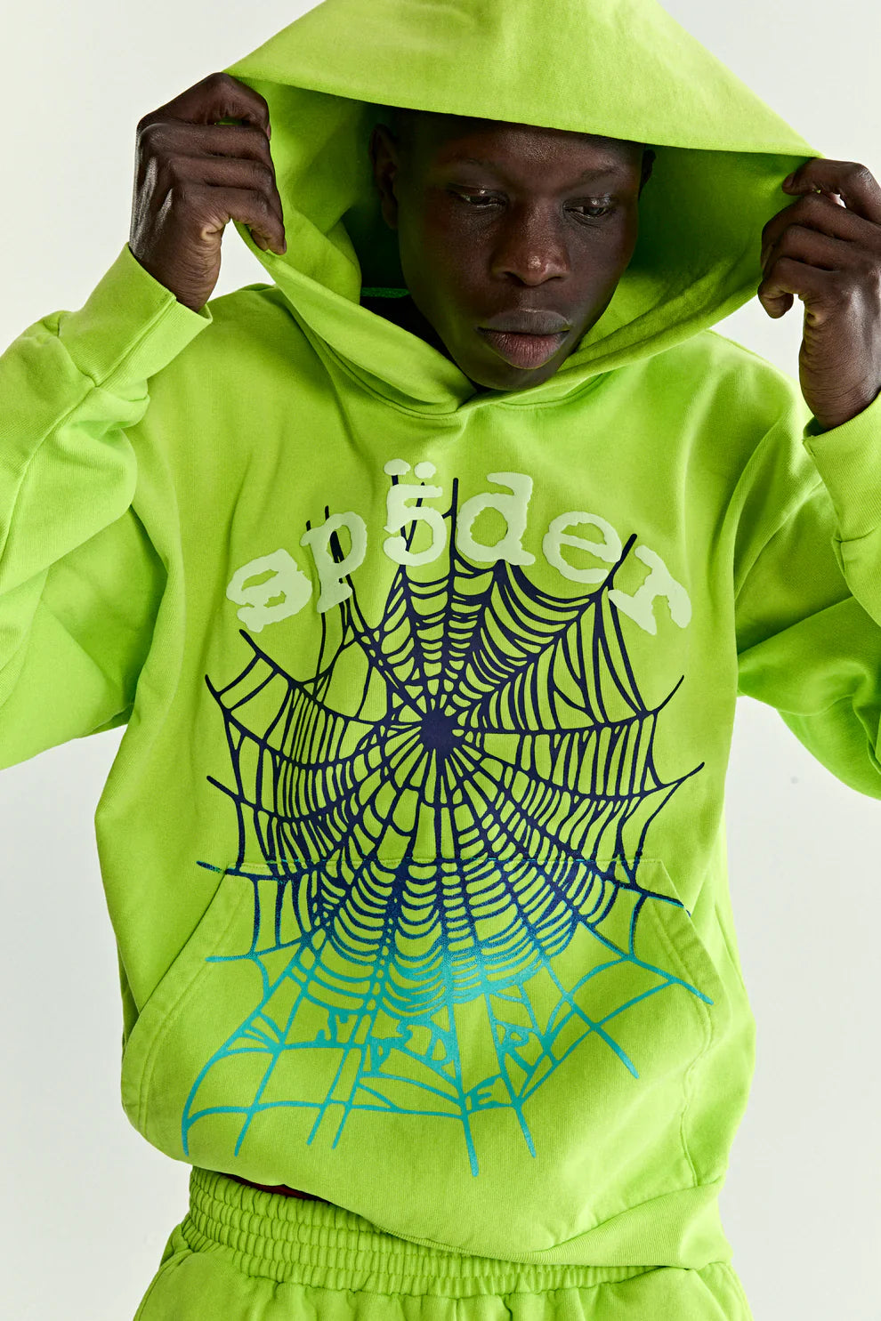 Wearing the Sp5der Web Graphic Hoodie – Neon Green 440GSM Heavyweight Streetwear, featuring bold black and blue spider web graphics, a person lifts the hood and looks downward.