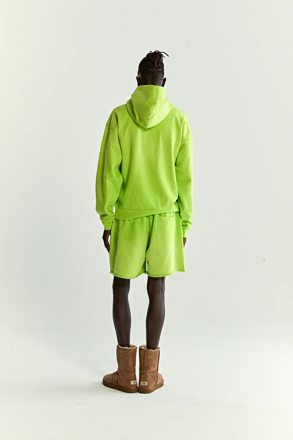 A person faces away, wearing a Sp5der Web Graphic Hoodie – Neon Green 440GSM Heavyweight Streetwear with matching shorts and brown boots, blending bold streetwear style against a plain light background.