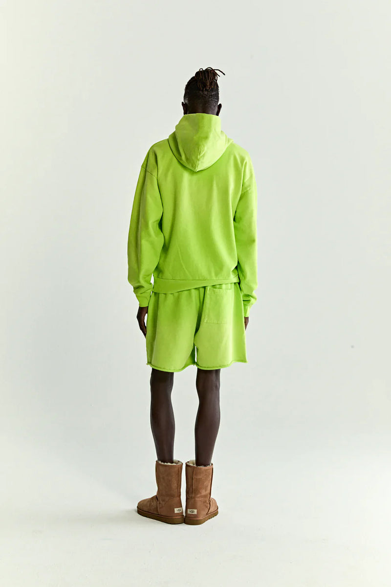 A person faces away, wearing a Sp5der Web Graphic Hoodie – Neon Green 440GSM Heavyweight Streetwear with matching shorts and brown boots, blending bold streetwear style against a plain light background.