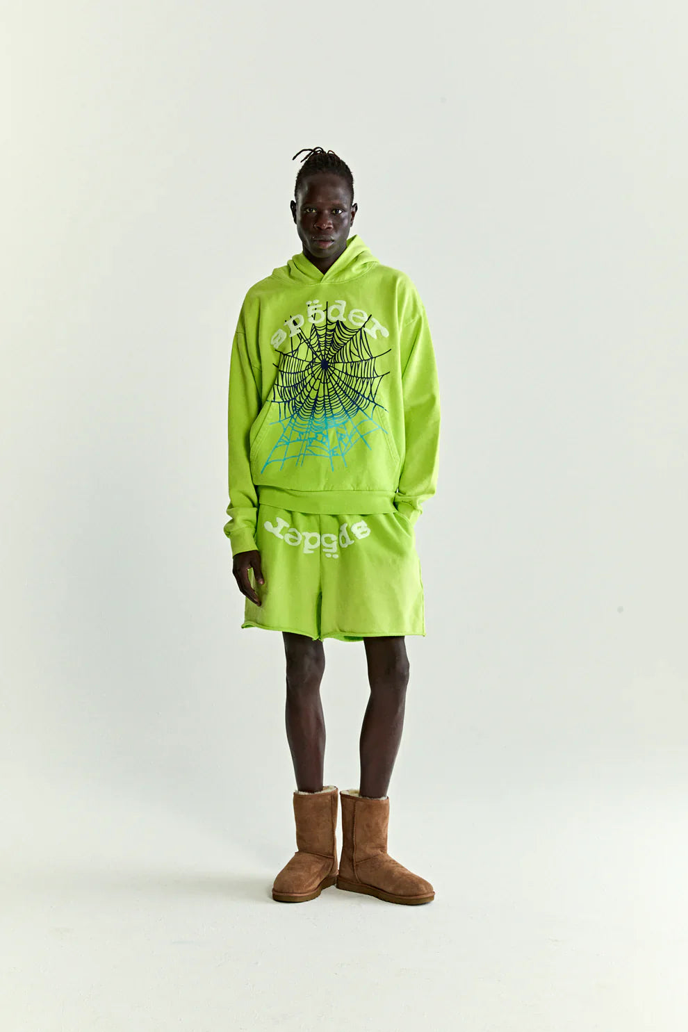 Wearing the Sp5der Web Graphic Hoodie – Neon Green 440GSM Heavyweight Streetwear with a black and white web graphic and matching shorts, a person stands against a plain background, finishing the look with brown boots.