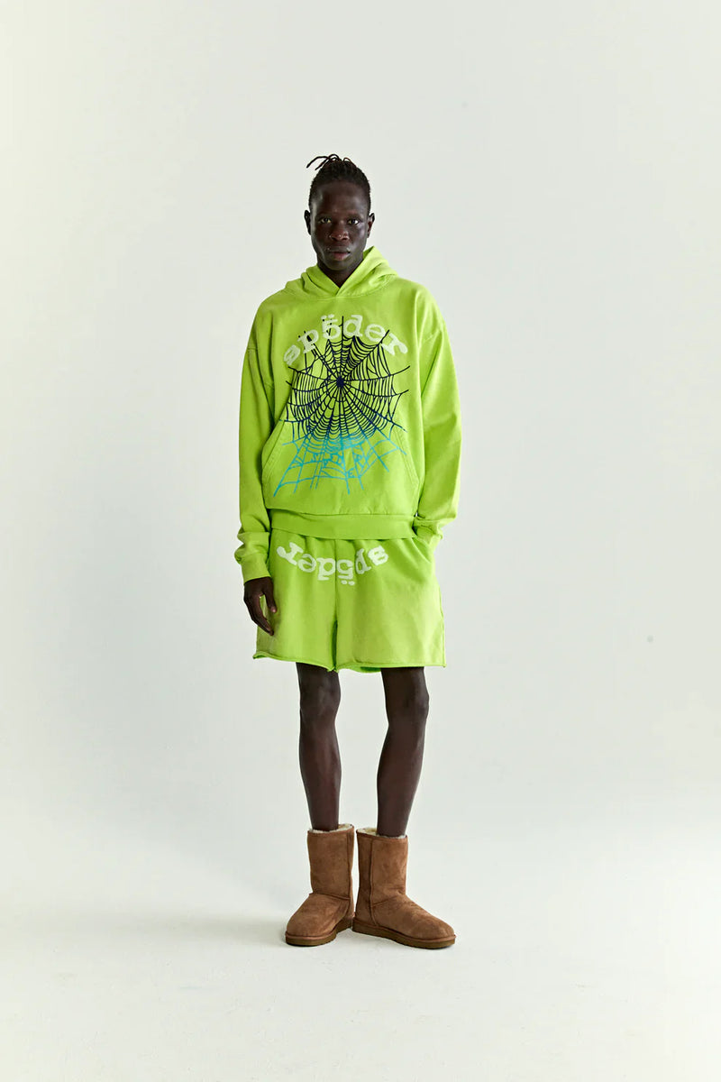 Wearing the Sp5der Web Graphic Hoodie – Neon Green 440GSM Heavyweight Streetwear with a black and white web graphic and matching shorts, a person stands against a plain background, finishing the look with brown boots.