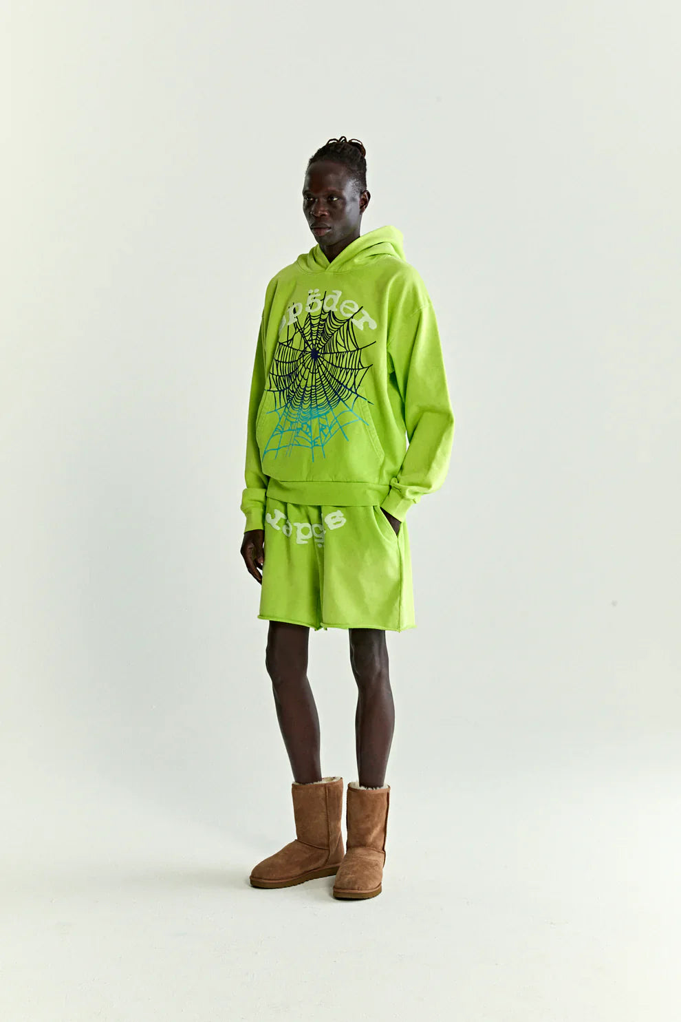 A person wears a Sp5der Web Graphic Hoodie – Neon Green 440GSM Heavyweight Streetwear with spiderweb graphics, paired with matching shorts featuring white text and brown suede boots, posed against a plain light background.