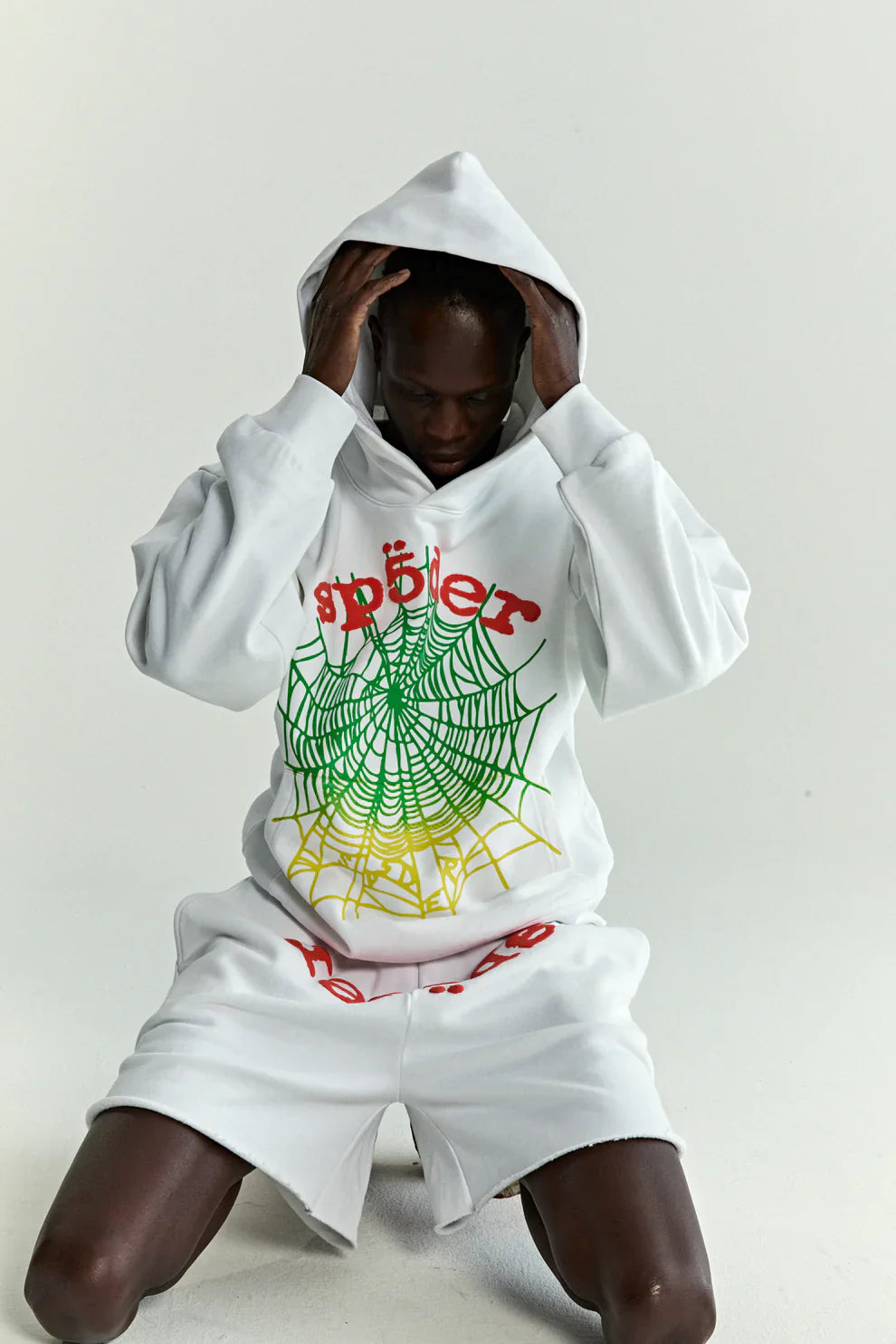 Wearing a Sp5der “Toxic Web” Hoodie – White & Neon Lime 440GSM Heavyweight Oversized Streetwear Pullover, a person in shorts crouches and holds the hood with both hands against a plain light background.