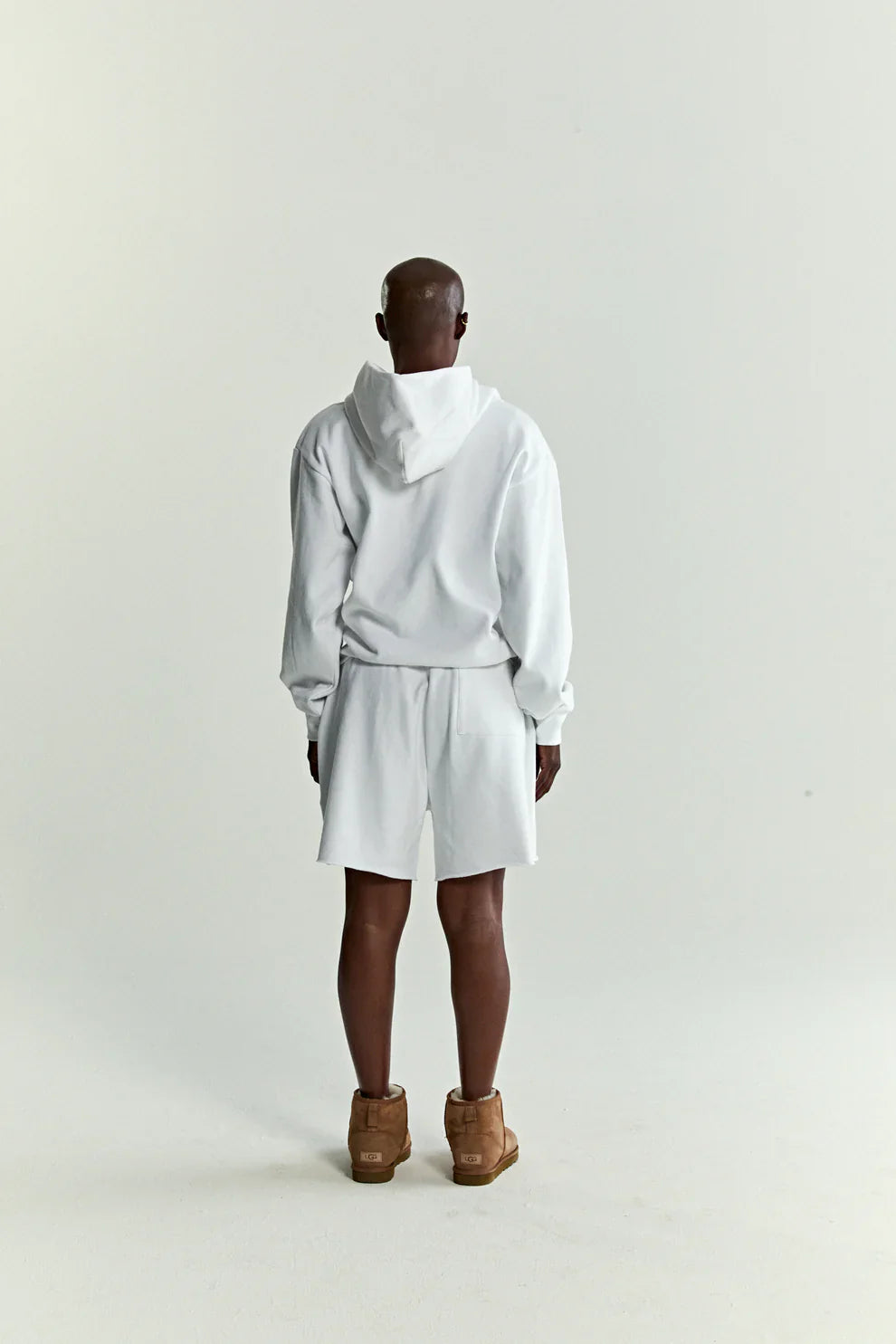 A person stands facing away in a Sp5der “Toxic Web” Hoodie – White & Neon Lime 440GSM Heavyweight Oversized Streetwear Pullover, white shorts, and tan shoes against a plain background—a cool tribute to Y2K street style.