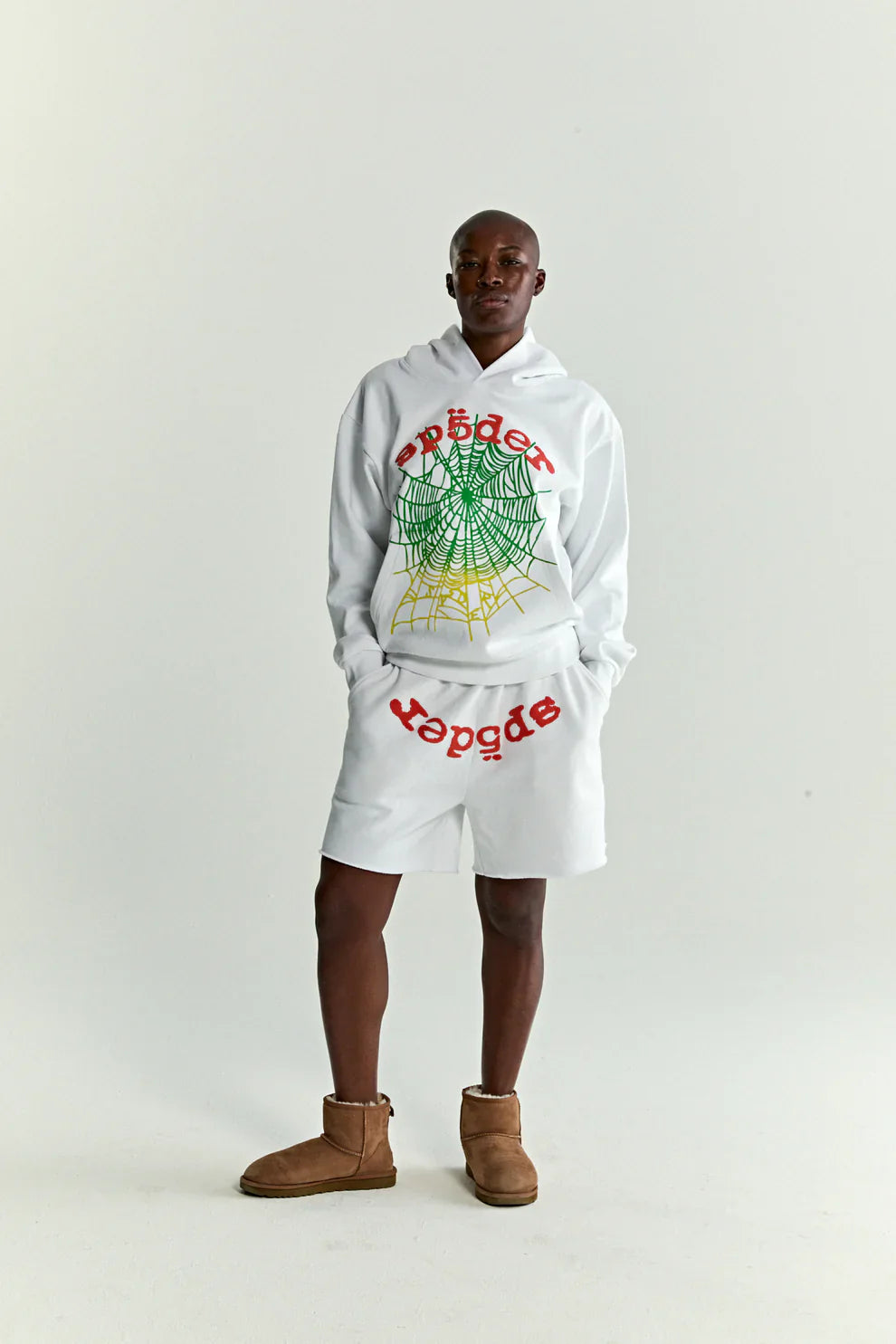 A person stands against a plain background in a white & neon lime Sp5der “Toxic Web” Hoodie, a 440GSM heavyweight oversized streetwear pullover with colorful spiderweb graphics, paired with matching shorts and brown ankle boots for a bold Y2K look.