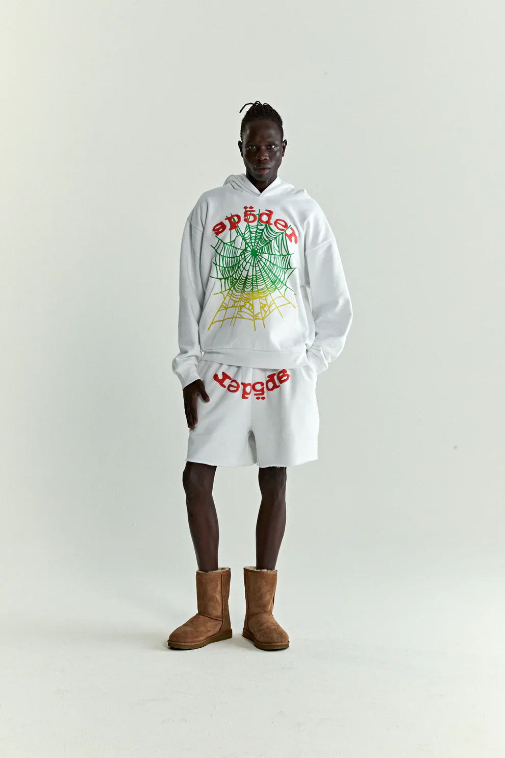 A person stands against a white background in the Sp5der “Toxic Web” Hoodie – White & Neon Lime 440GSM Heavyweight Oversized Streetwear Pullover, paired with white shorts featuring red text and brown boots for a bold Y2K streetwear look.
