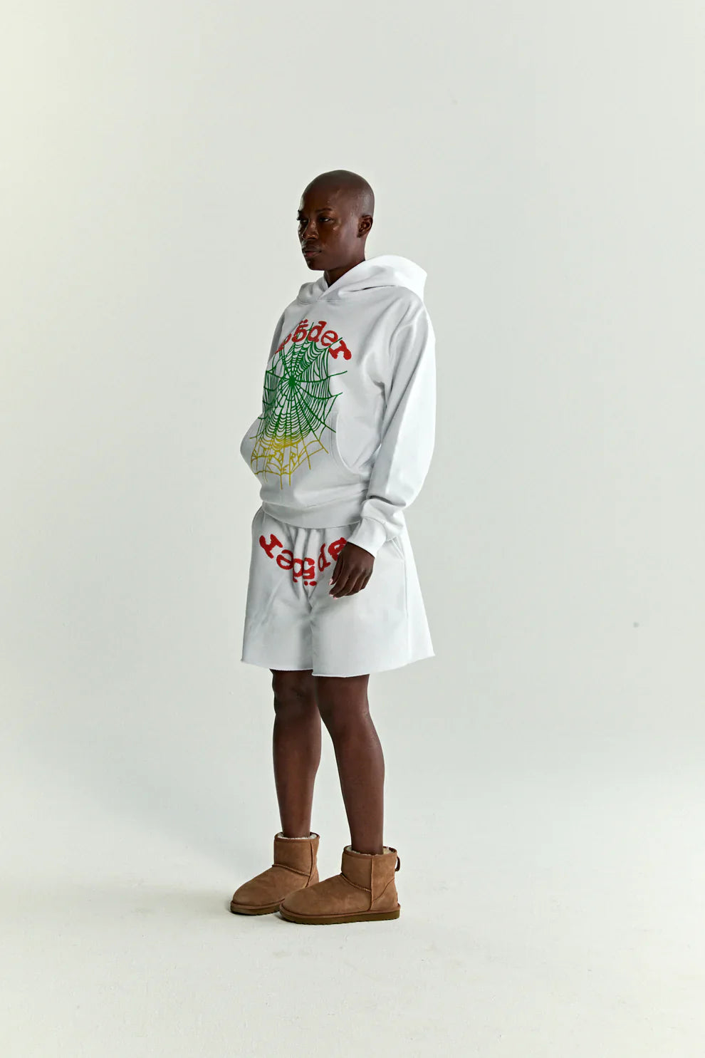 Someone stands against a plain background in a Sp5der “Toxic Web” Hoodie – White & Neon Lime 440GSM heavyweight oversized streetwear pullover, paired with a white knee-length skirt and light brown ankle boots, hands in pocket and facing slightly sideways.