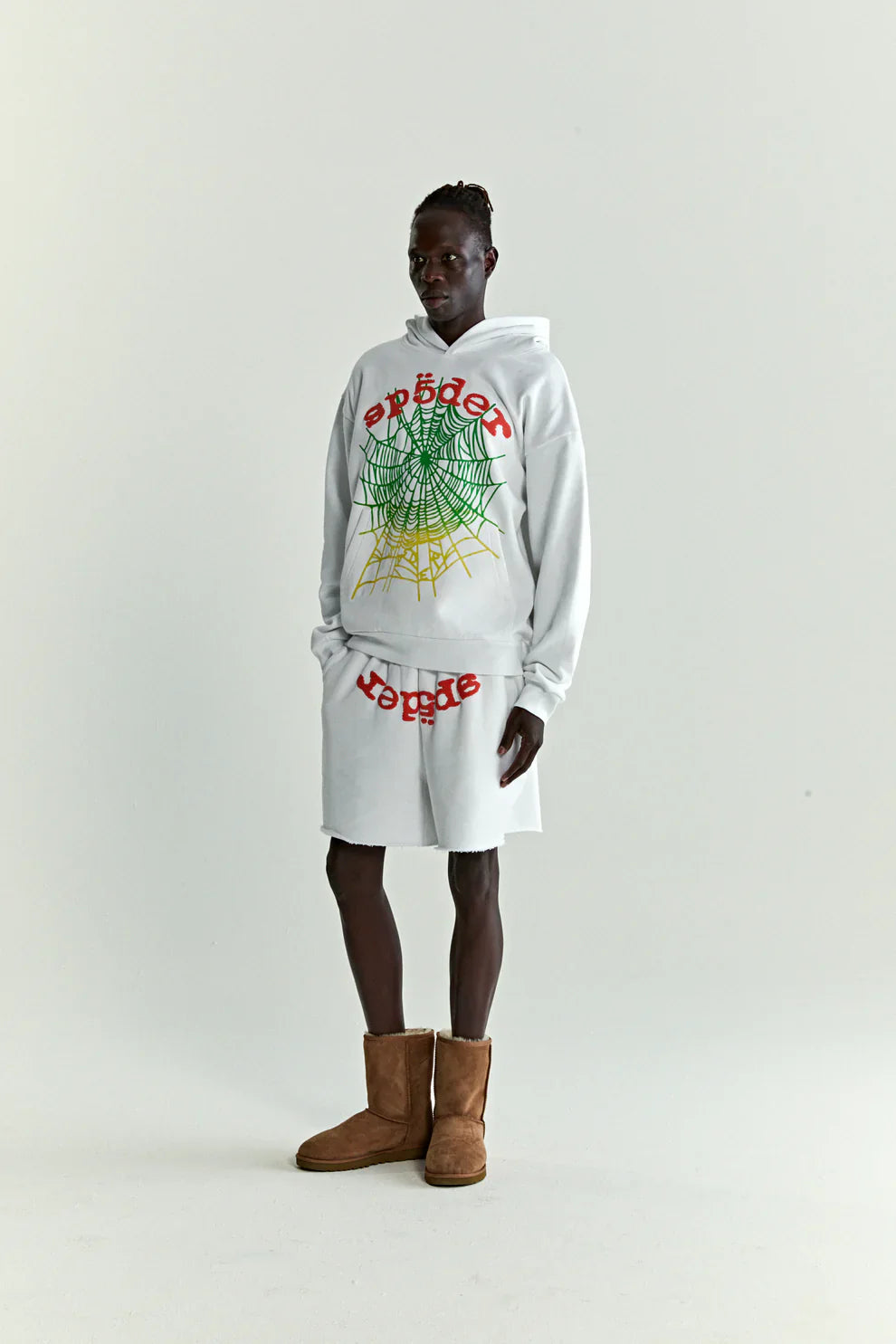 A person stands against a plain background wearing a Sp5der “Toxic Web” Hoodie in white & neon lime, a 440GSM heavyweight oversized streetwear pullover, paired with matching shorts with colorful graphics and tan suede boots for a bold Y2K vibe.