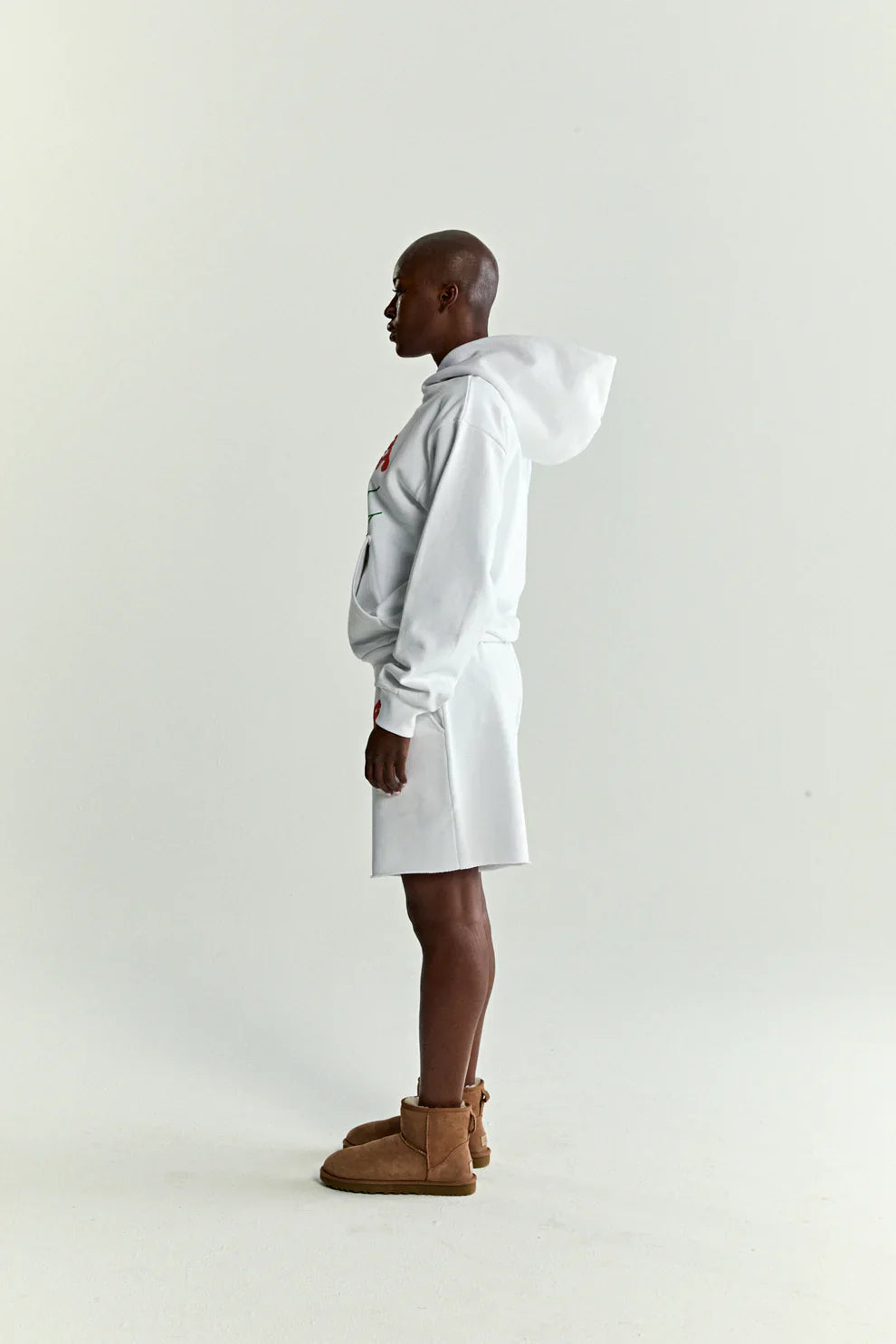 A person stands in profile facing left, wearing a Sp5der “Toxic Web” Hoodie – White & Neon Lime 440GSM Heavyweight Oversized Streetwear Pullover, white shorts, and brown ankle boots against a plain light background.