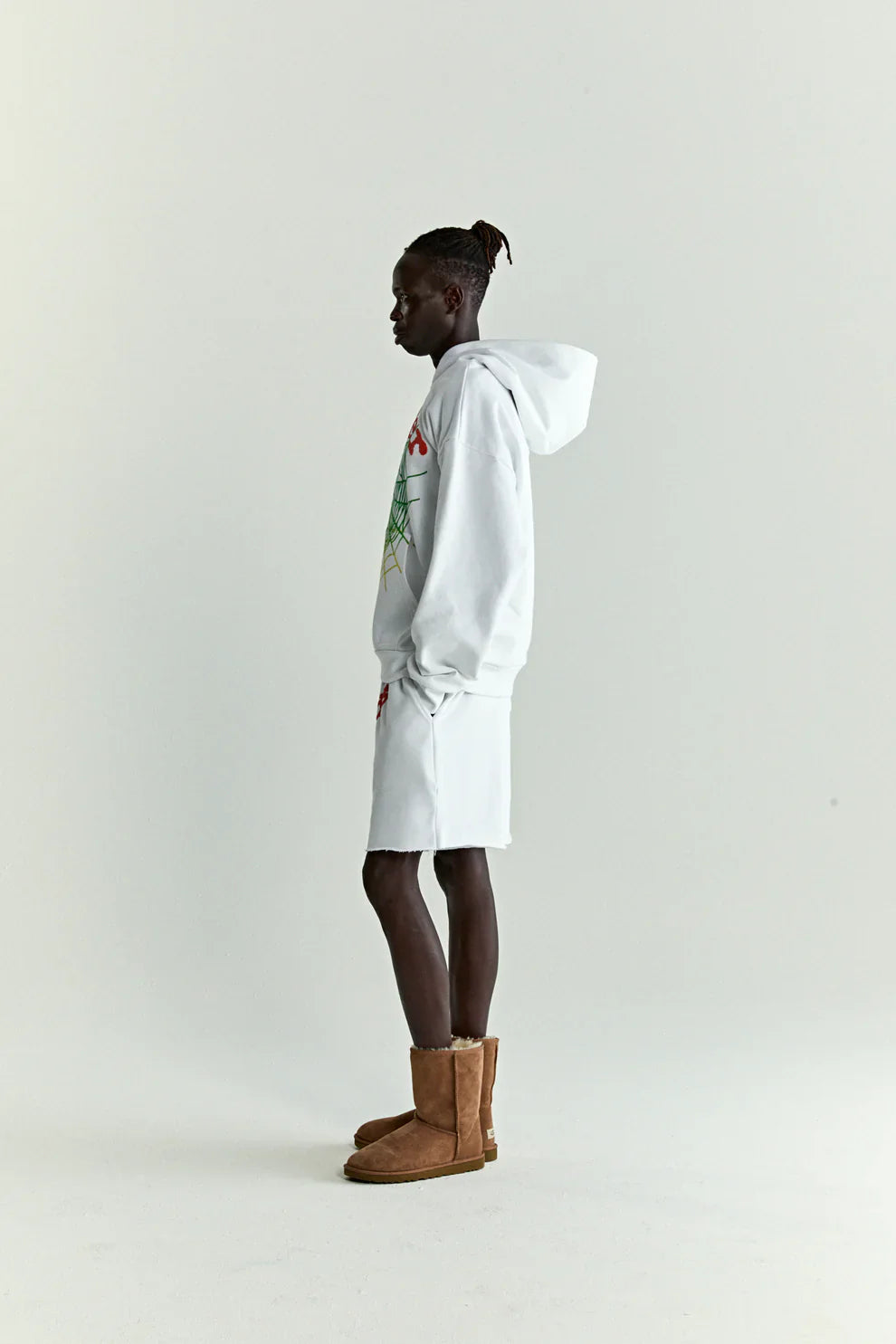 A person in profile against a plain background wears a white & neon lime Sp5der “Toxic Web” Hoodie, an oversized 440GSM heavyweight streetwear pullover, paired with white shorts and brown boots. Their short twists pulled back evoke Y2K street style.