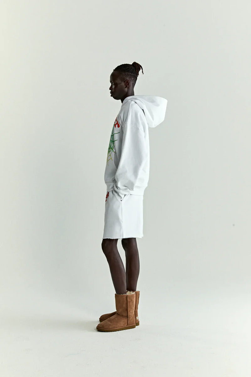 A person in profile against a plain background wears a white & neon lime Sp5der “Toxic Web” Hoodie, an oversized 440GSM heavyweight streetwear pullover, paired with white shorts and brown boots. Their short twists pulled back evoke Y2K street style.