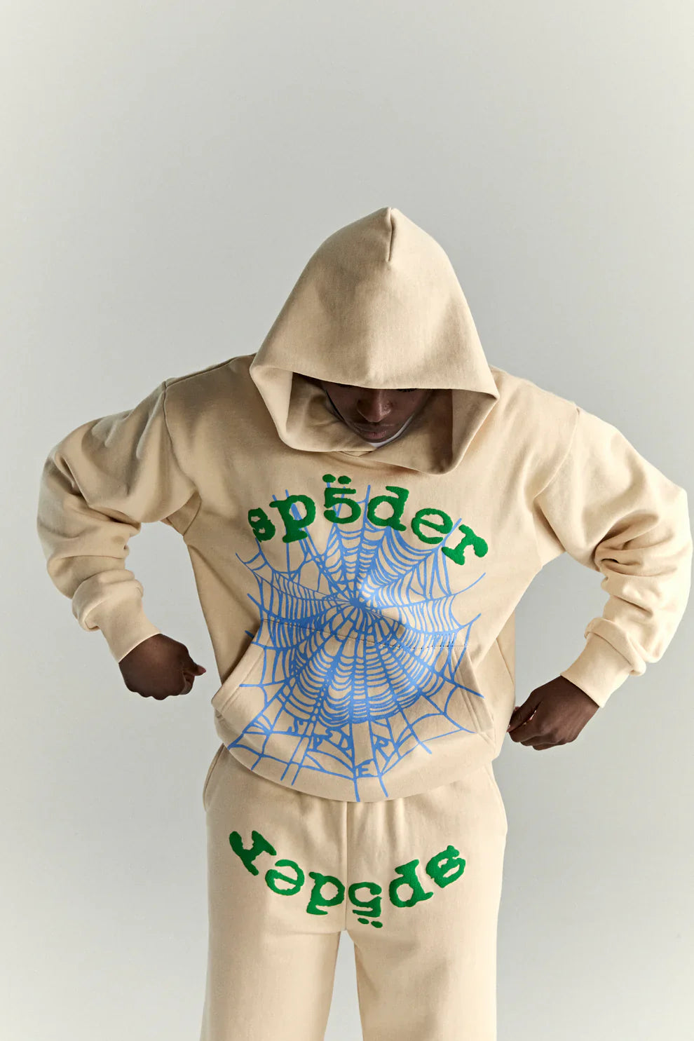 Wearing an oversized Sp5der “Sky Web” Hoodie in cream, blue, and green (440GSM), featuring a blue spider web and green Sp5der text, this Y2K streetwear look is finished with the hood up and head lowered.