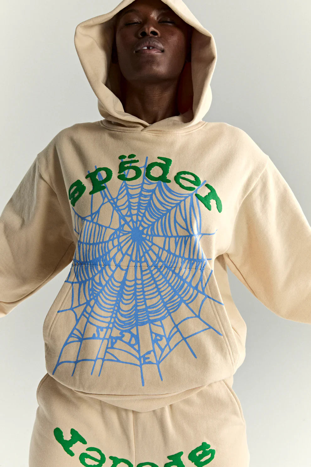A person in a Sp5der “Sky Web” Hoodie – Cream/Blue/Green 440GSM oversized streetwear pullover with bold green text and a large blue spiderweb graphic, matching pants, posing Y2K style with arms out and eyes closed.