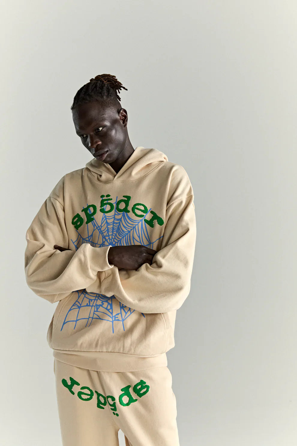 A person in the Sp5der “Sky Web” Hoodie (Cream/Blue/Green, 440GSM oversized pullover) with matching pants, featuring green "sp5der" lettering and a blue web graphic, poses against a light gray background—Y2K streetwear style.