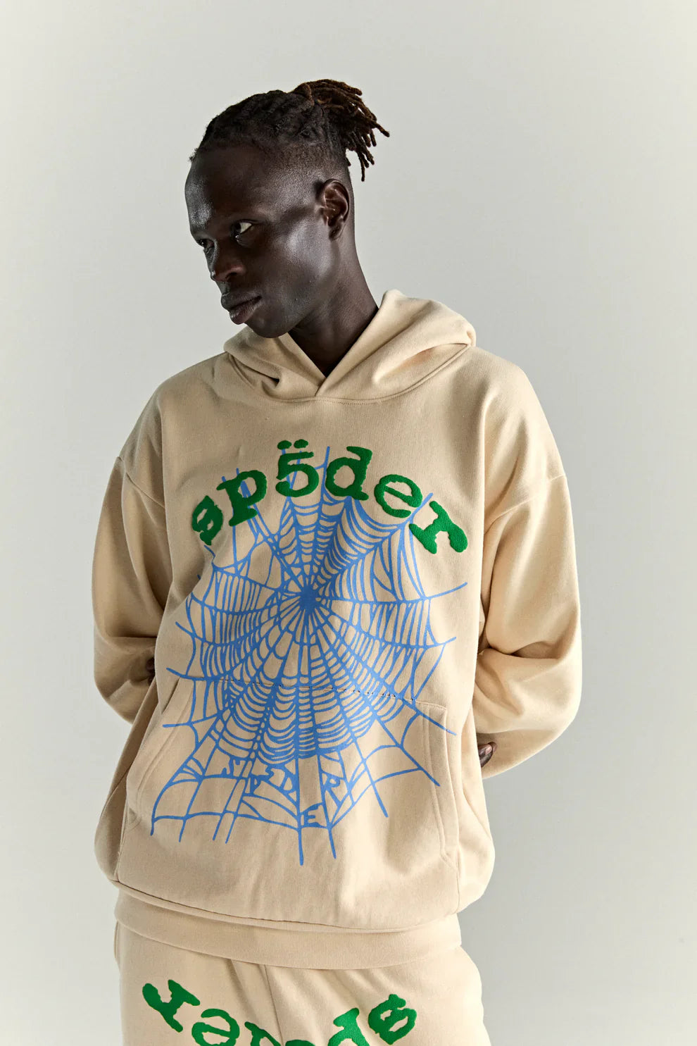 Wearing the Sp5der “Sky Web” Hoodie in cream with blue and green details, a person stands relaxed against a light background, hands behind their back, showcasing oversized Y2K-inspired streetwear style.