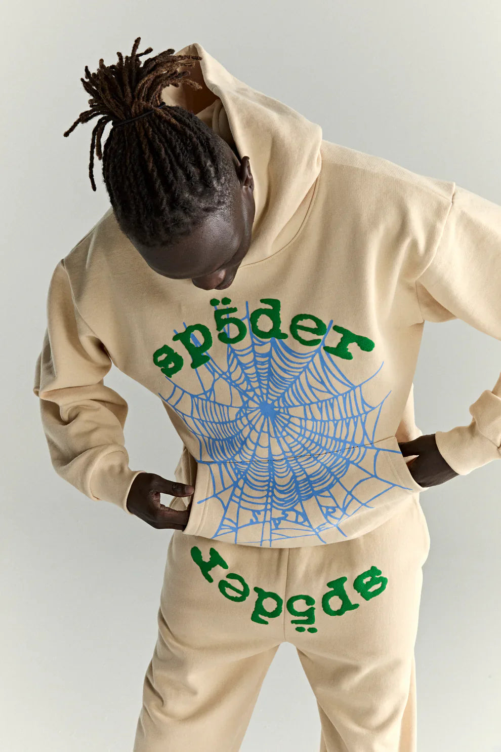 Wearing a Sp5der “Sky Web” Hoodie—Cream/Blue/Green 440GSM oversized streetwear pullover—a person showcases bold Y2K style, with green Sp5der text and a large blue spiderweb graphic, looking down at the design.