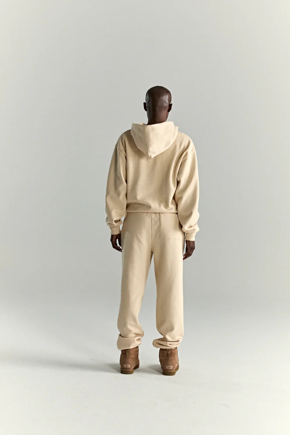 A person stands facing away from the camera wearing a Sp5der “Sky Web” Hoodie in cream/blue/green (440GSM oversized streetwear pullover) and matching sweatpants, with brown shoes, against a plain light background.