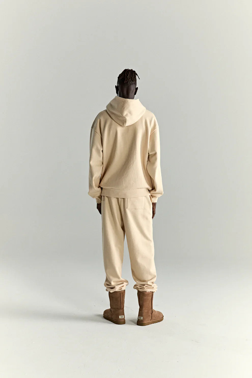 A person faces away from the camera wearing the Sp5der “Sky Web” Hoodie in cream/blue/green (440GSM oversized streetwear pullover), matching sweatpants, and brown boots against a plain light gray background.