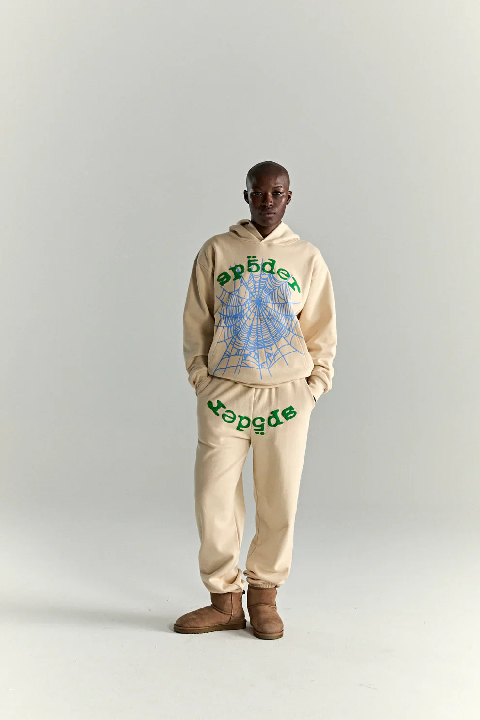 A person stands against a plain light background, wearing a Sp5der “Sky Web” Hoodie in cream with blue and green graphics, 440GSM heavyweight cotton fleece, styled in Y2K streetwear fashion with matching sweatpants and brown slip-on boots.