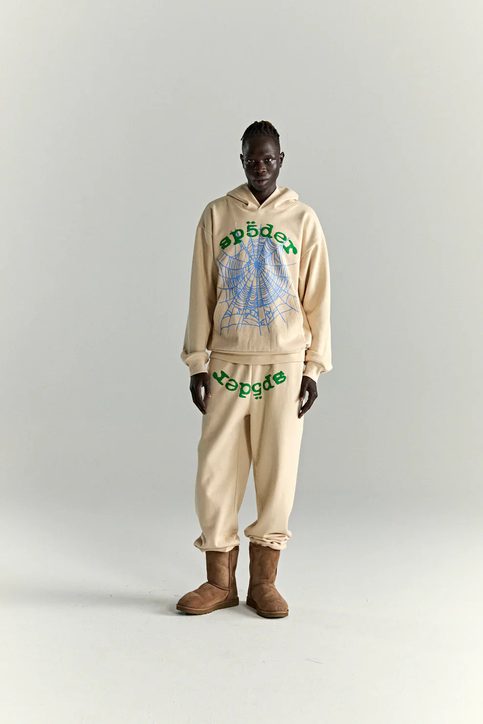 A person stands against a light background, wearing a Sp5der "Sky Web" Hoodie in cream with green text and blue web graphics, plus matching sweatpants—both heavyweight cotton fleece—styled with brown boots for a Y2K streetwear vibe.