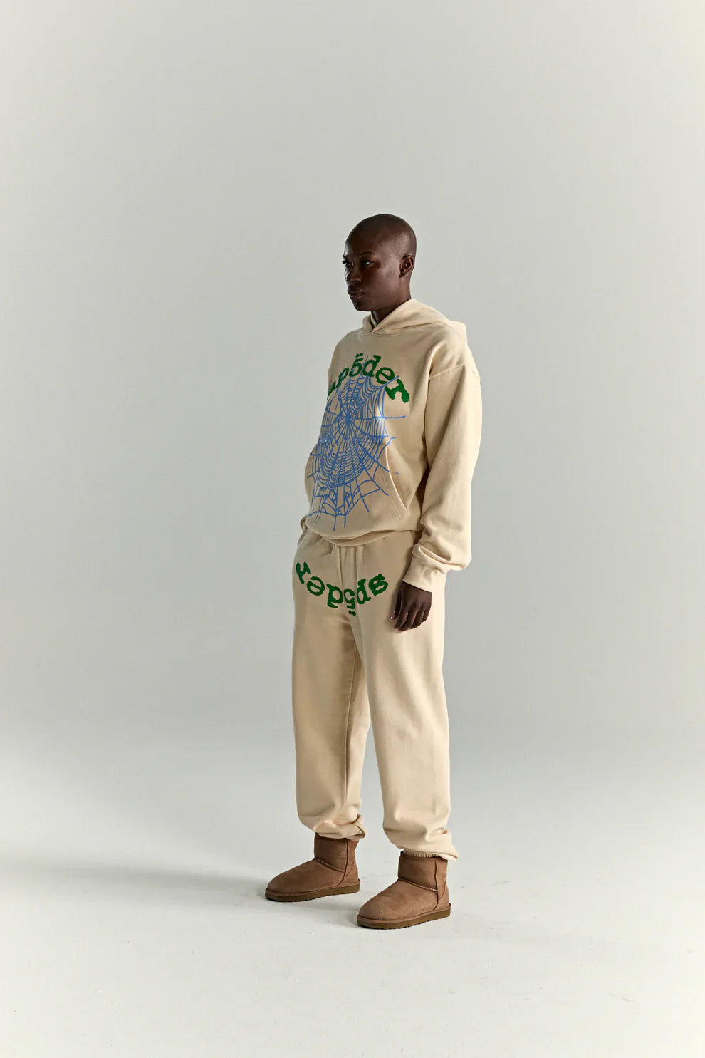 A person stands in profile wearing the Sp5der “Sky Web” Hoodie – Cream/Blue/Green, a 440GSM oversized streetwear pullover, paired with matching pants and brown slippers against a plain light gray background, channeling Y2K vibes.