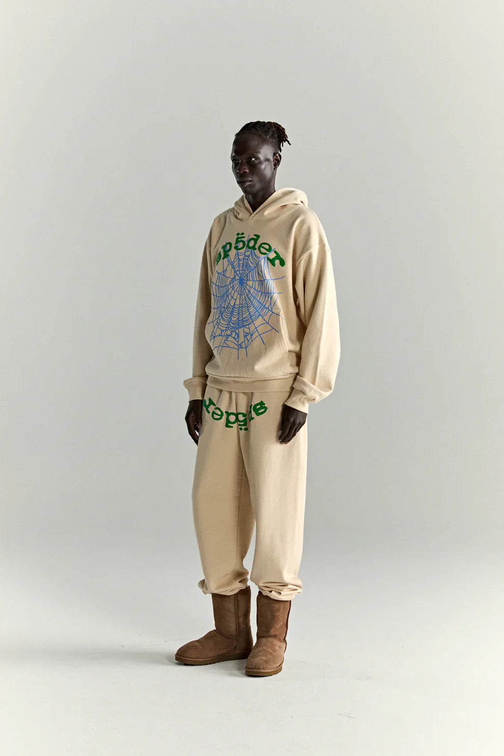A person stands against a plain background in a Sp5der “Sky Web” Hoodie—cream with blue and green accents, 440GSM heavyweight cotton fleece—paired with matching sweatpants and brown boots for a Y2K streetwear vibe.