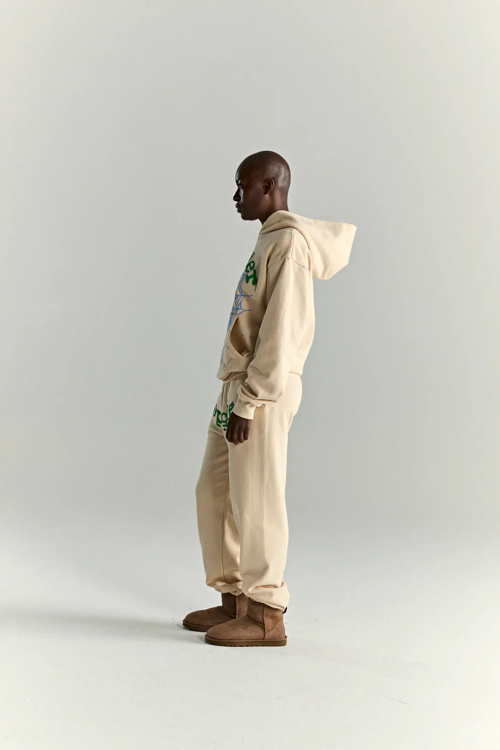 A person in a Sp5der “Sky Web” Hoodie – Cream / Blue / Green 440GSM Oversized Streetwear Pullover stands in profile against a plain background, wearing brown boots and sweatpants, exuding effortless Y2K streetwear with heavyweight fleece comfort.