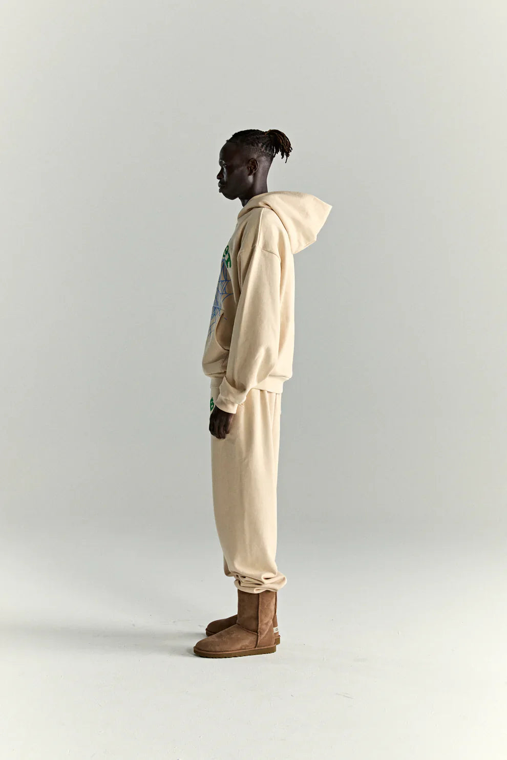 A person in profile wears a Sp5der “Sky Web” Hoodie – Cream / Blue / Green 440GSM oversized streetwear pullover, paired with matching sweatpants and brown boots. The hoodie features colorful graphics, creating a Y2K vibe with short twist hair.