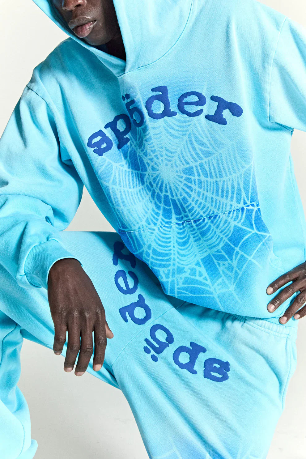 Someone wearing the Sp5der “Blue Venom” Web Hoodie—an oversized streetwear pullover in sky blue fade, made from 440GSM heavyweight cotton fleece, featuring bold "spider" lettering and a large spiderweb graphic on the front.