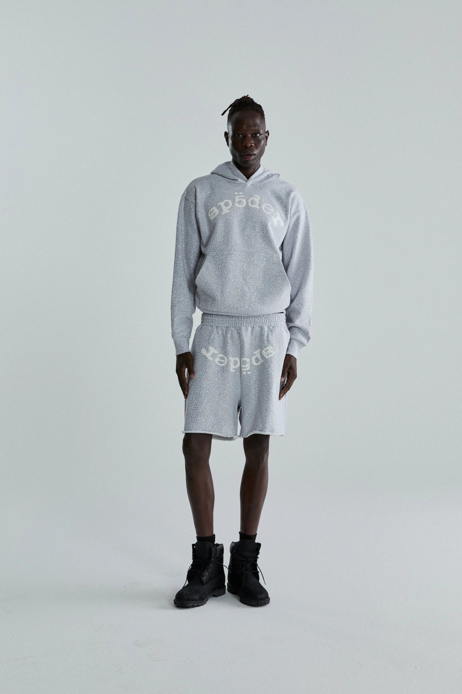 A person stands against a plain light background wearing a Grey Sp5der Rhinestone VVS Hoodie 440GSM – Grey Crystal Sp5der Hoodie All-Over Rhinestones and matching shorts, both light gray with “speed” printed upside down, paired with black high-top shoes.