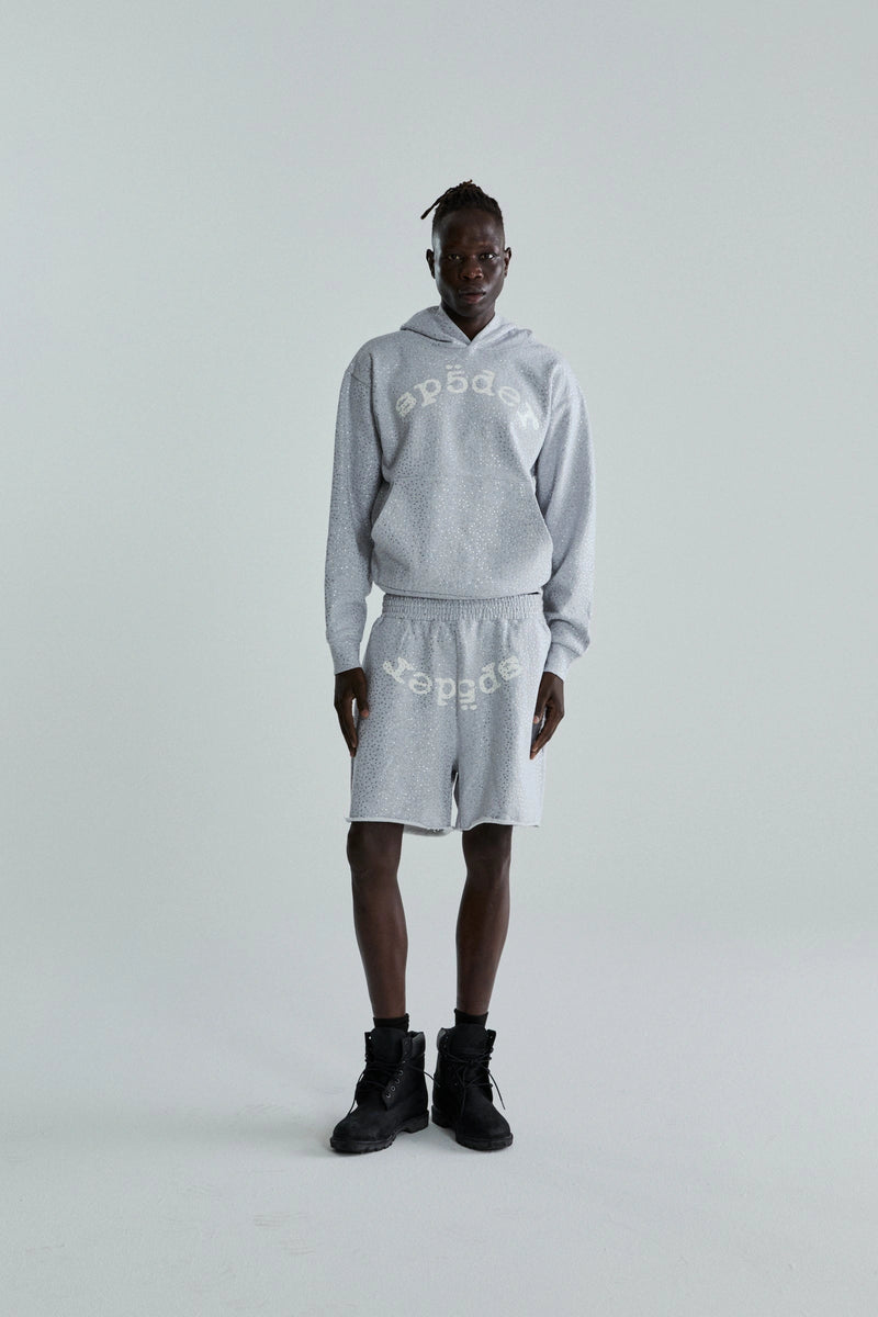 A person stands against a plain light background wearing a Grey Sp5der Rhinestone VVS Hoodie 440GSM – Grey Crystal Sp5der Hoodie All-Over Rhinestones and matching shorts, both light gray with “speed” printed upside down, paired with black high-top shoes.