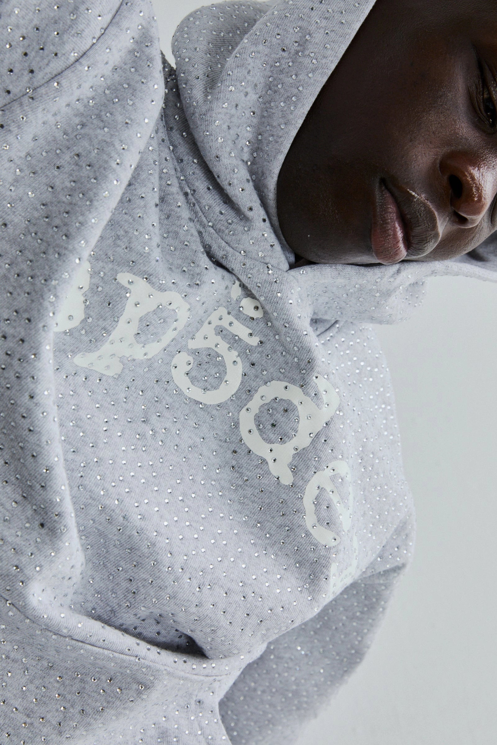A person wearing the Grey Sp5der Rhinestone VVS Hoodie 440GSM, featuring all-over rhinestones and a raised white design, looks down with the hood up.