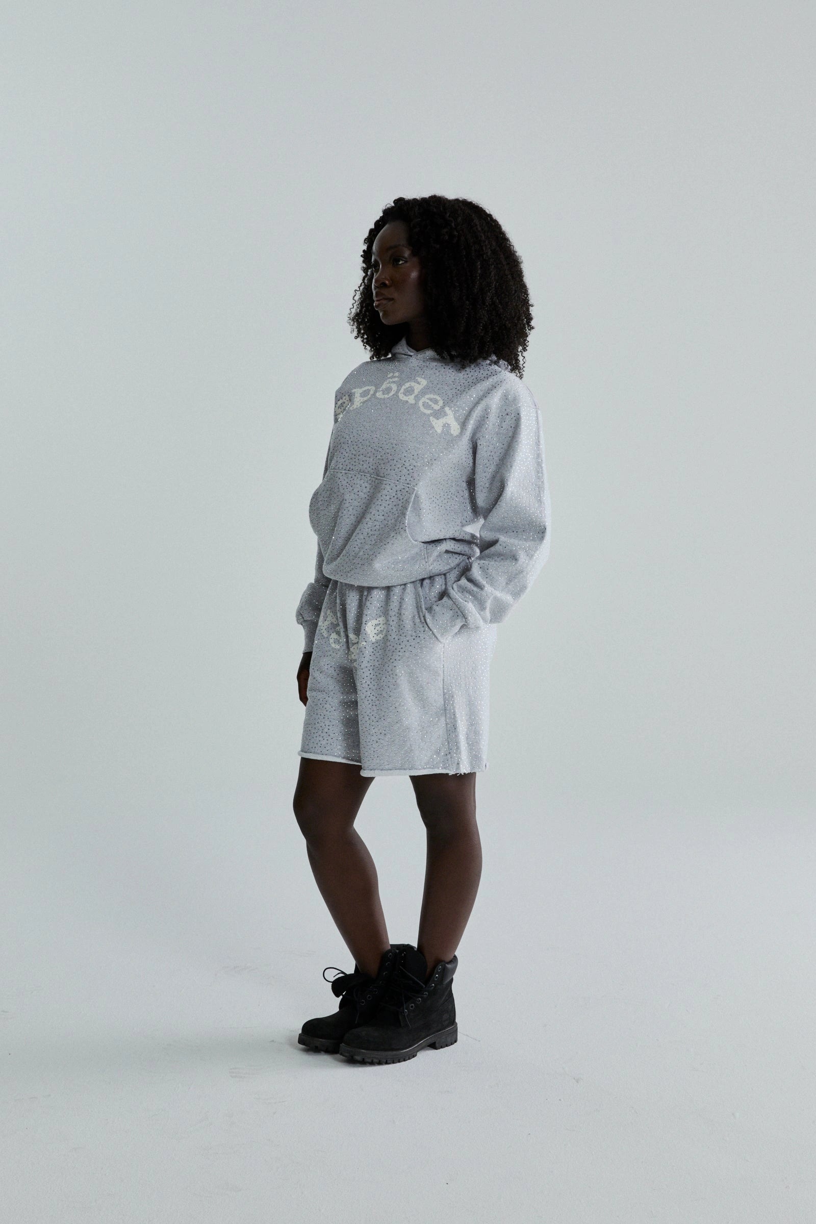 A woman with curly hair stands sideways, wearing a Grey Sp5der Rhinestone VVS Hoodie 440GSM – Grey Crystal Sp5der Hoodie All-Over Rhinestones styled oversized with matching shorts and black ankle boots, against a plain light background.