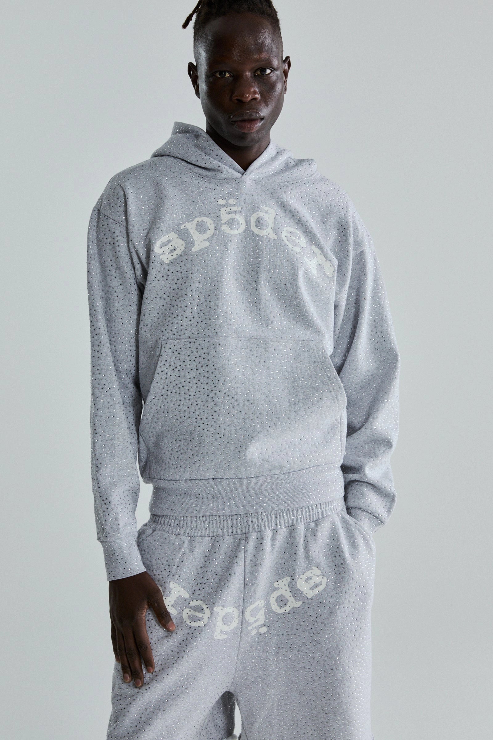 A person stands against a plain background wearing the Grey Sp5der Rhinestone VVS Hoodie 440GSM, featuring all-over rhinestones and "sp5der" in white on light gray fabric. The look is completed with matching sweatpants.