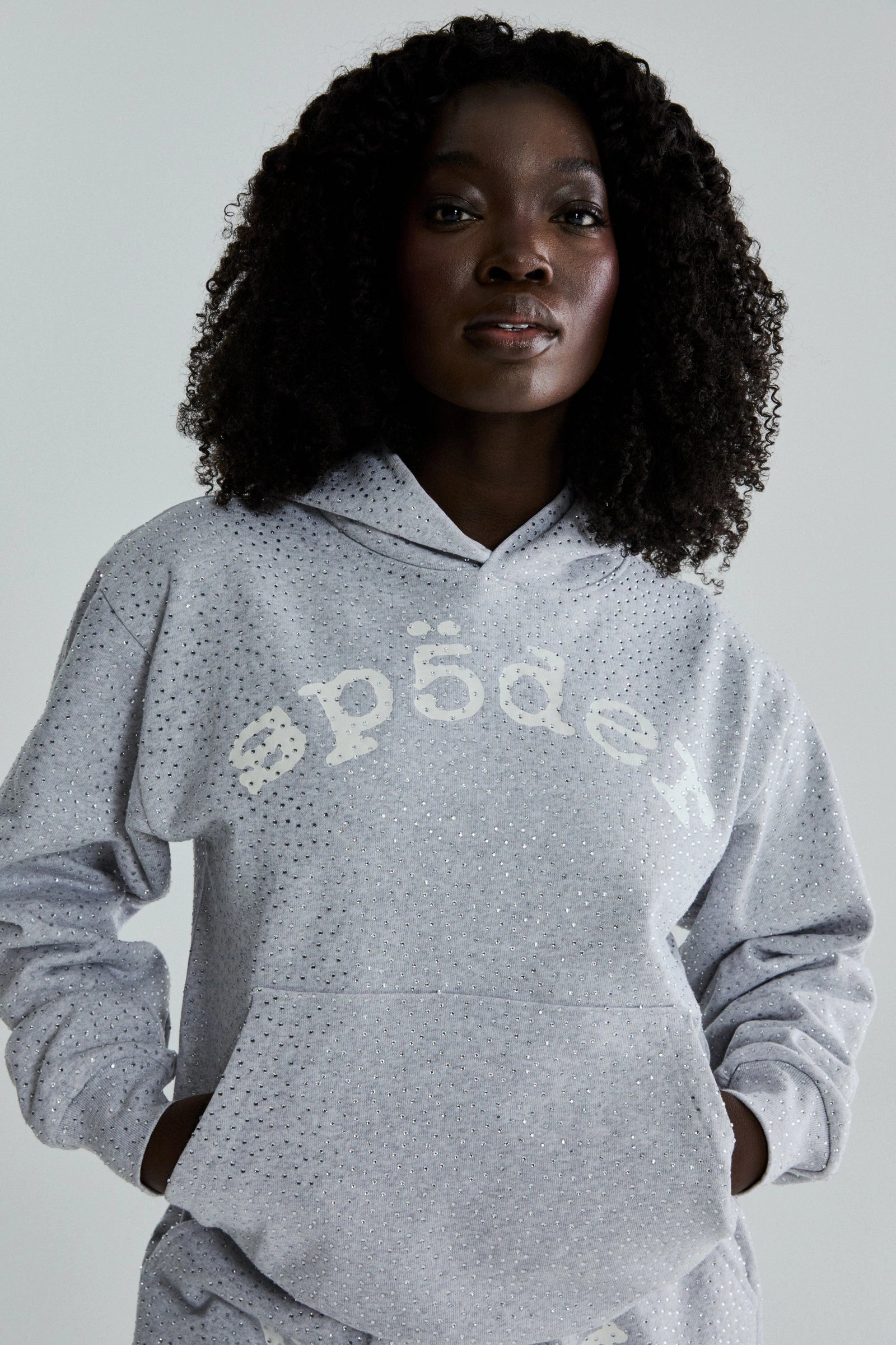 A woman with curly hair confidently wears the Grey Sp5der Rhinestone VVS Hoodie 440GSM—a light gray hooded sweatshirt featuring all-over rhinestones and stylized white "sp5der" lettering, hands tucked in the front pocket.
