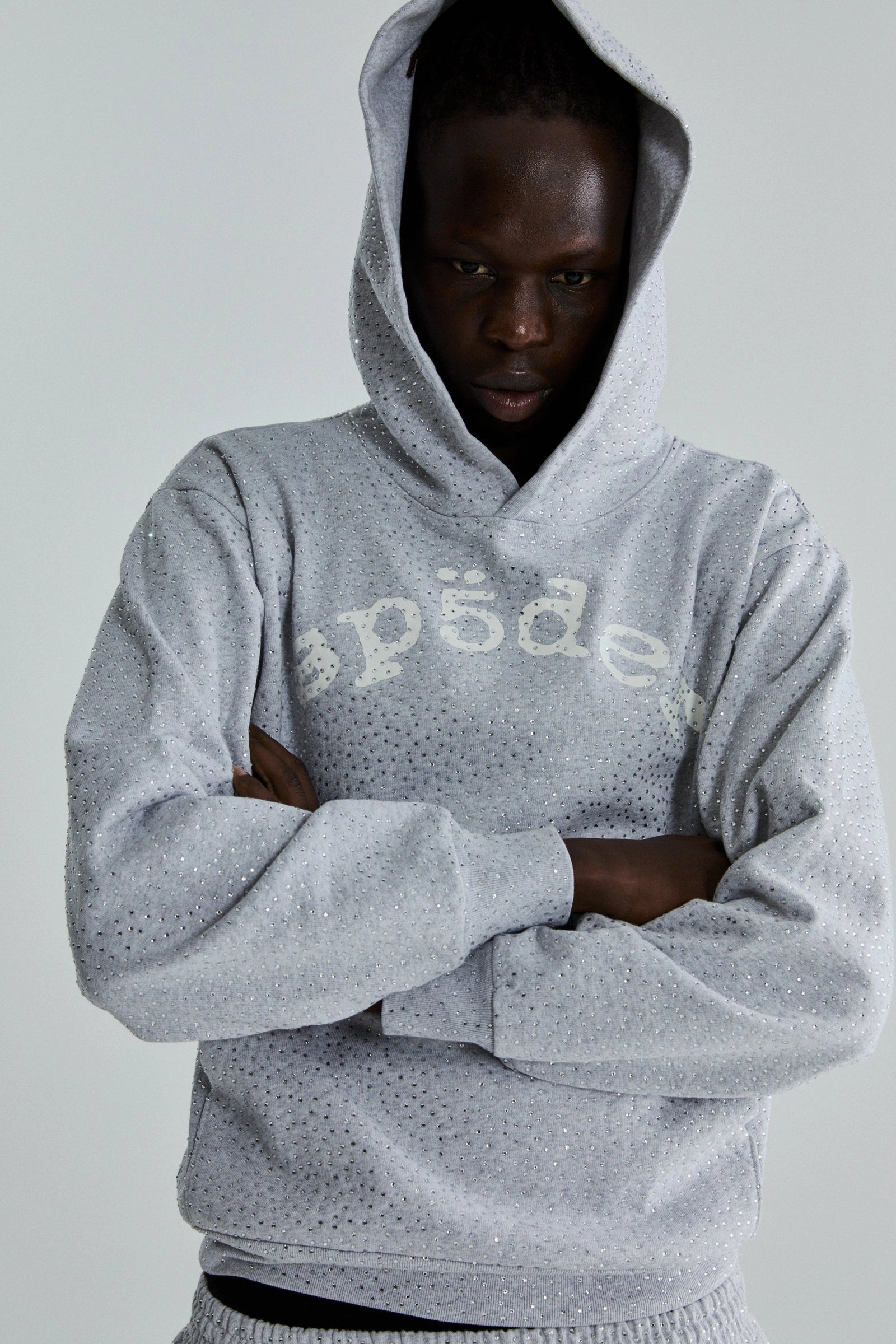 A person in the Grey Sp5der Rhinestone VVS Hoodie 440GSM—an all-over rhinestone grey hoodie—stands with arms crossed, looking downward.