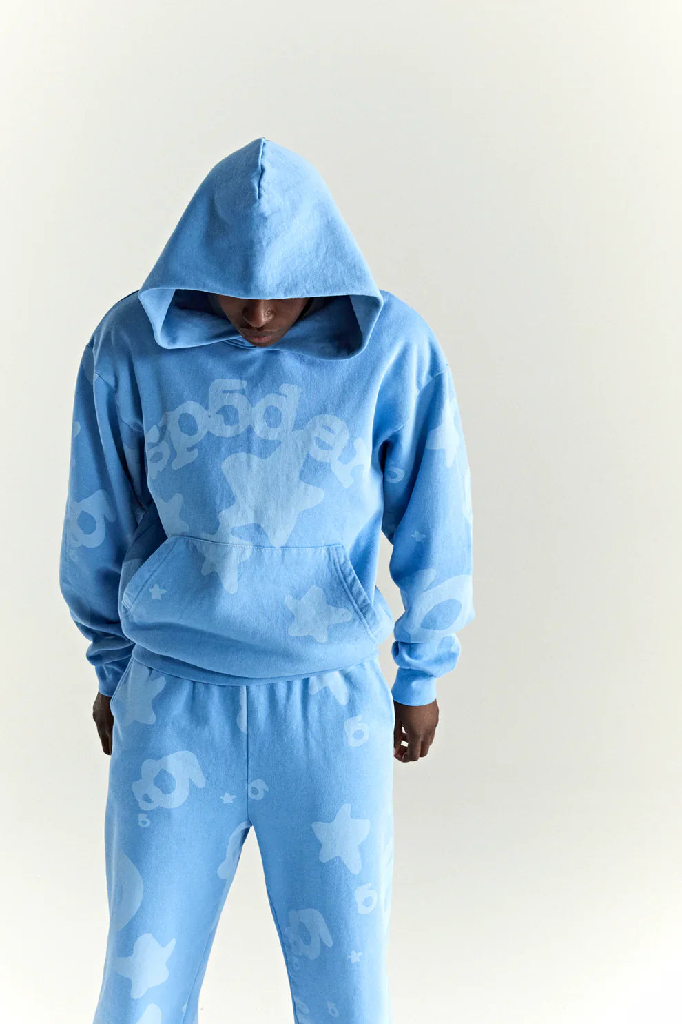 A person wearing the Sp5der Star Hoodie – Baby Blue 440GSM Heavyweight Oversized Streetwear Pullover and matching sweatpants looks down, with the hood covering most of their head, set against a plain light background.