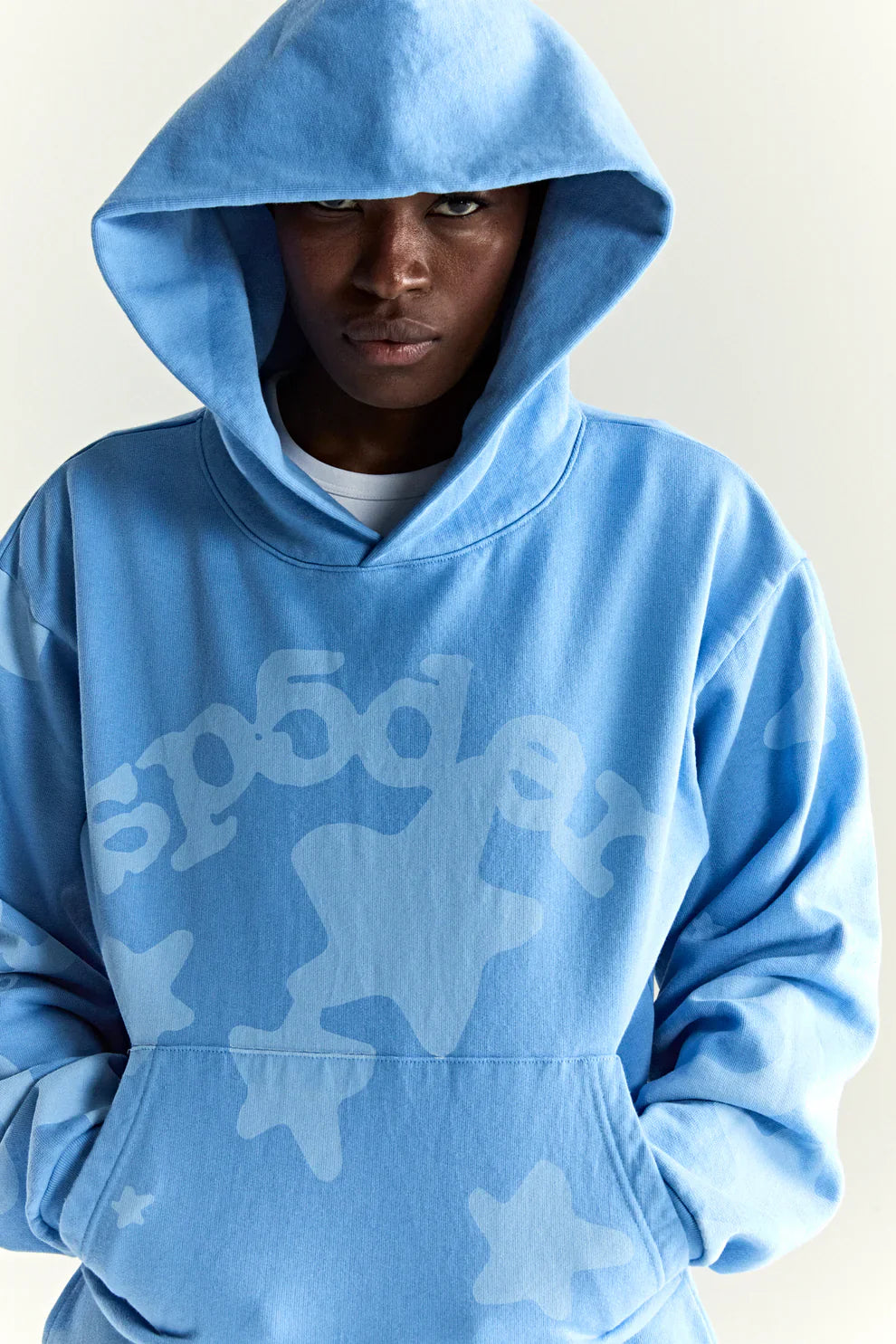 Wearing the Sp5der Star Hoodie – Baby Blue 440GSM Heavyweight Oversized Streetwear Pullover with white star designs and the hood up, a person stands against a plain background, showcasing bold Y2K-inspired streetwear style.