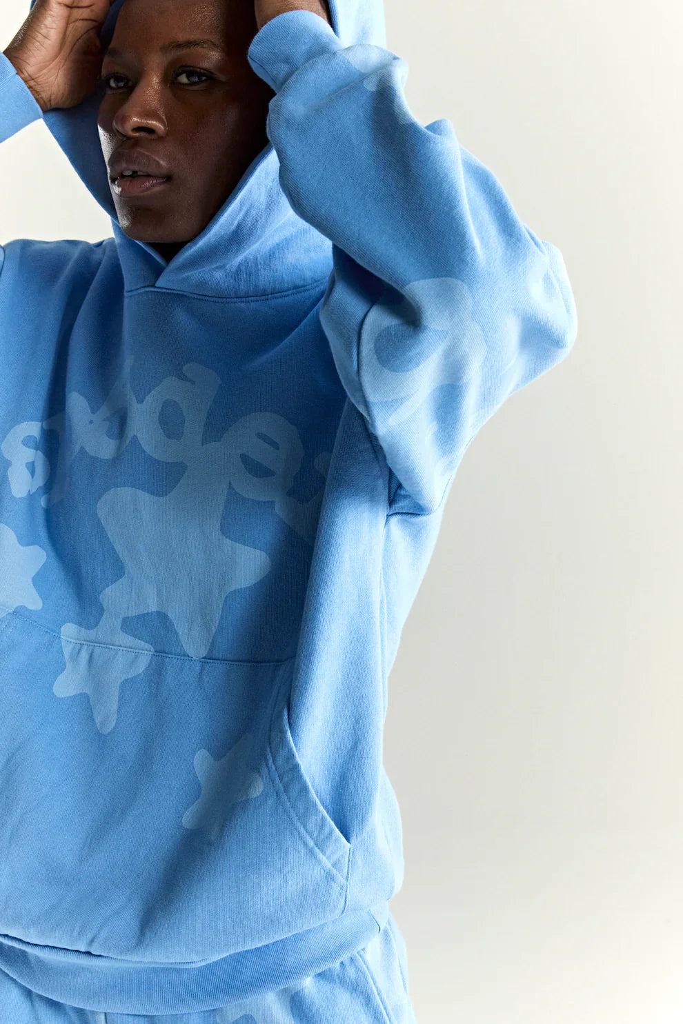 A person wears the Sp5der Star Hoodie – Baby Blue 440GSM Heavyweight Oversized Streetwear Pullover with matching pants, pulling the hood up and posing against a plain backdrop—delivering iconic Y2K streetwear style in premium cotton fleece.