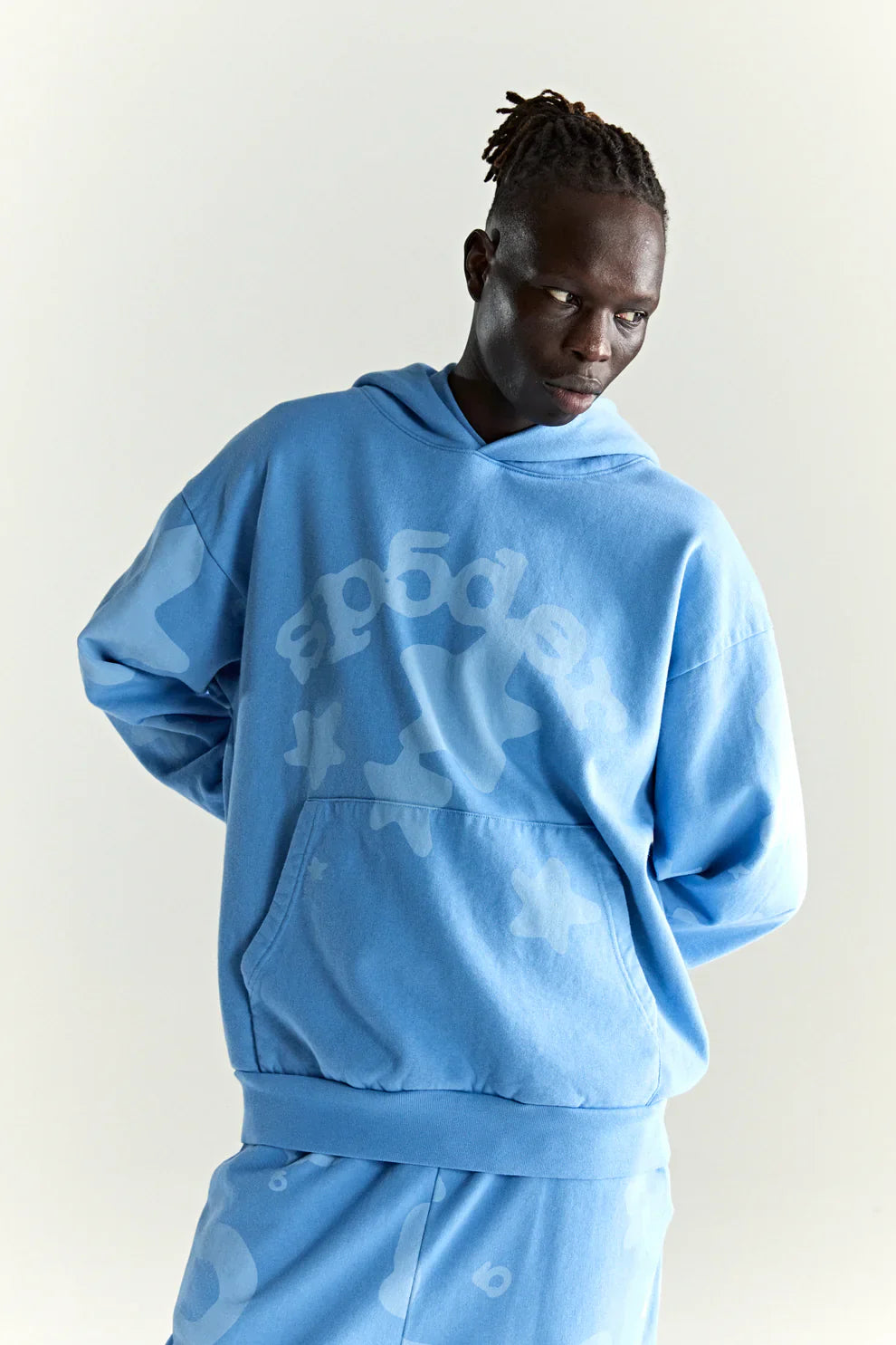 Wearing the Sp5der Star Hoodie – Baby Blue 440GSM Heavyweight Oversized Streetwear Pullover, a person channels Y2K streetwear vibes against a plain background, looking left with hands behind their back in an easy, loose fit.