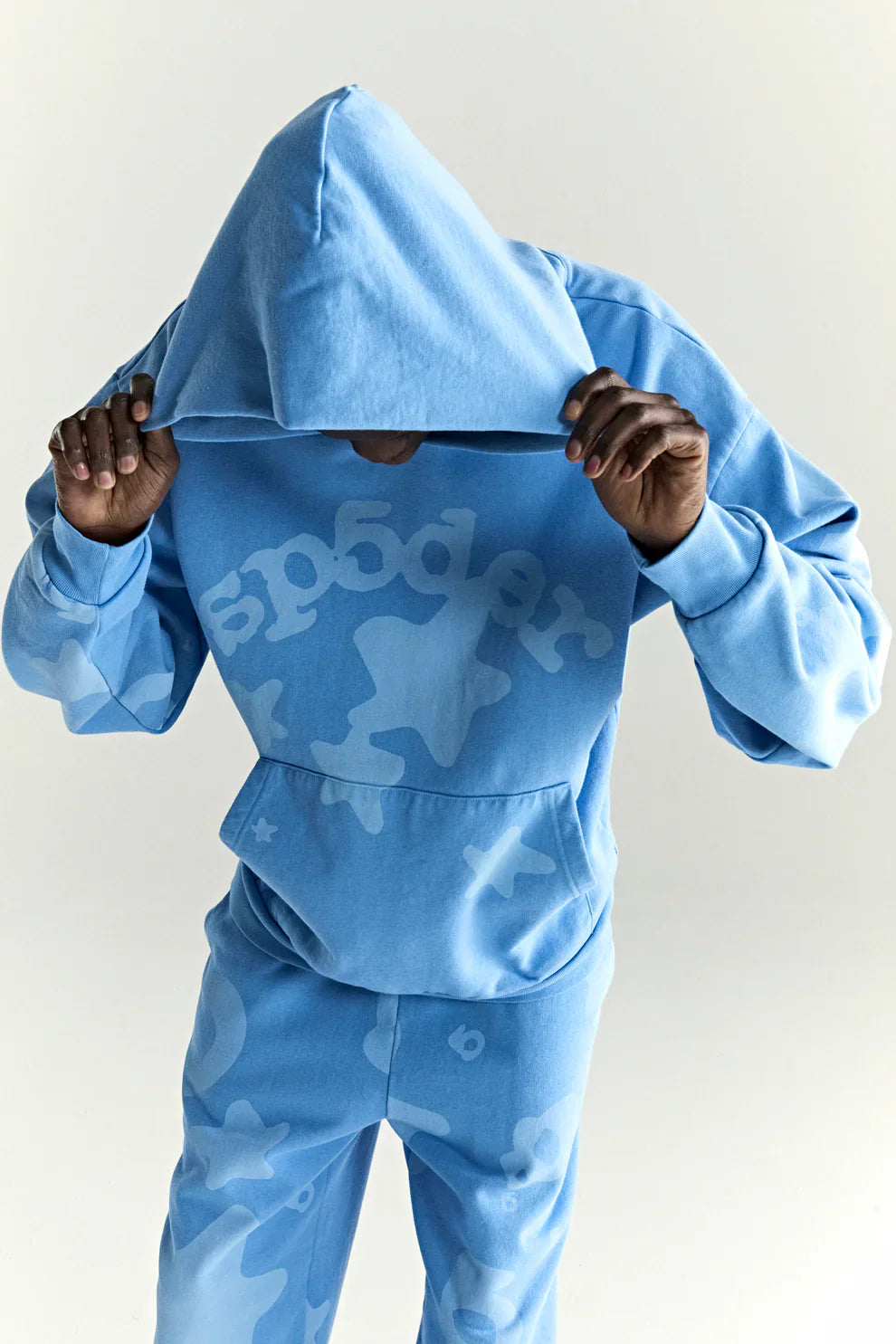 Wearing the Sp5der Star Hoodie in Baby Blue—an oversized 440GSM heavyweight streetwear pullover—a person pulls up the hood, its abstract white patterns and Y2K vibe standing out against a plain light background.