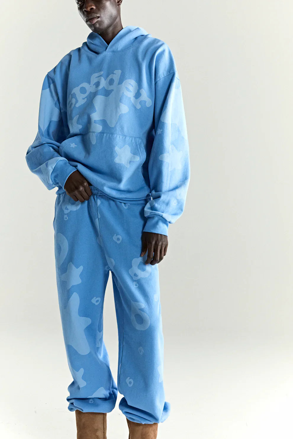 A person stands wearing a loose-fitting Sp5der Star Hoodie – Baby Blue 440GSM Heavyweight Oversized Streetwear Pullover and matching sweatpants, both with an abstract pattern and "Sp5der" on the chest, paired with tan boots against a plain light background.