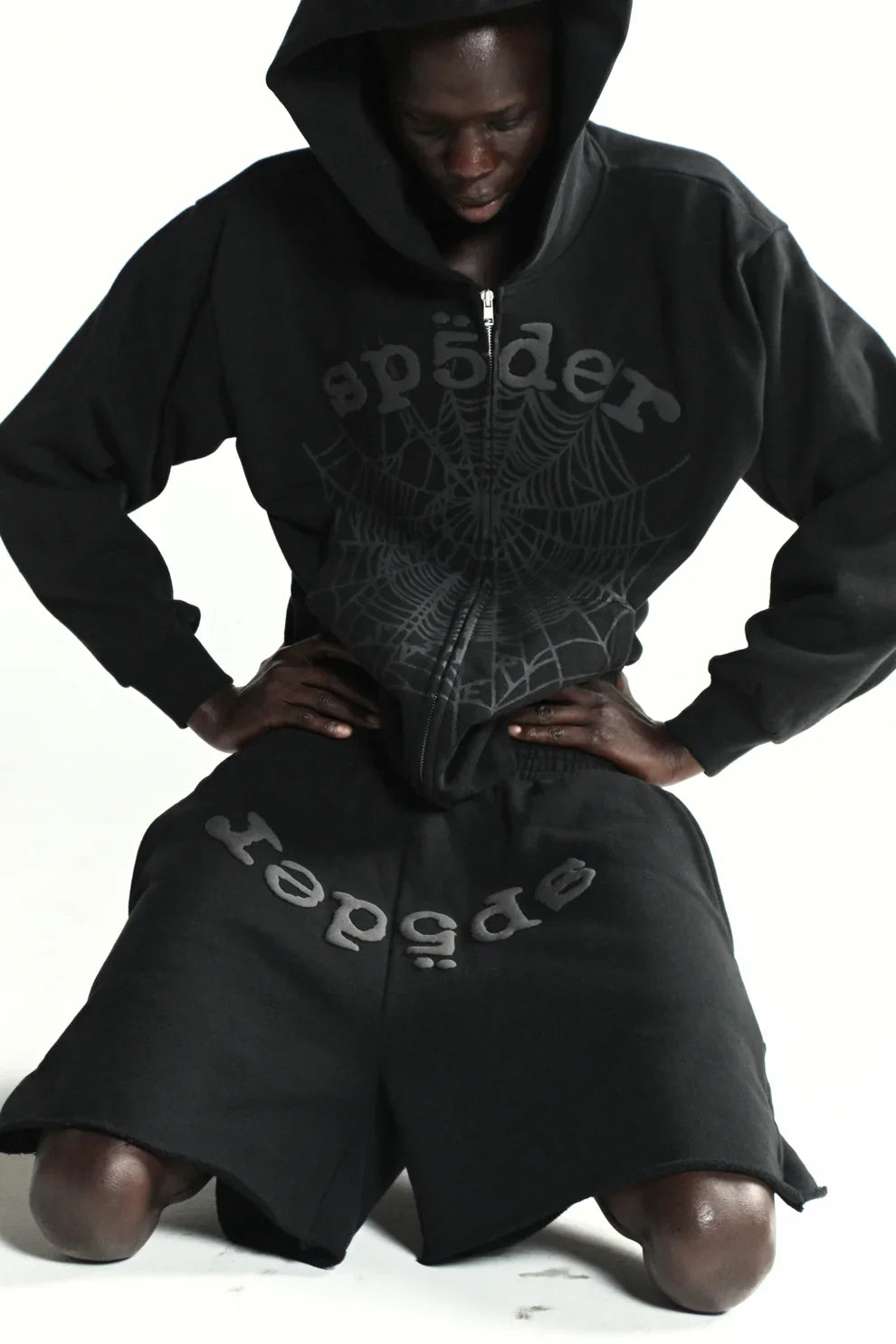 A person kneels on the floor against a white background, wearing the Sp5der Tonal Web Zip Hoodie – Black 440GSM Heavyweight Streetwear with the hood up, highlighting its bold tonal spider web graphic and street style.