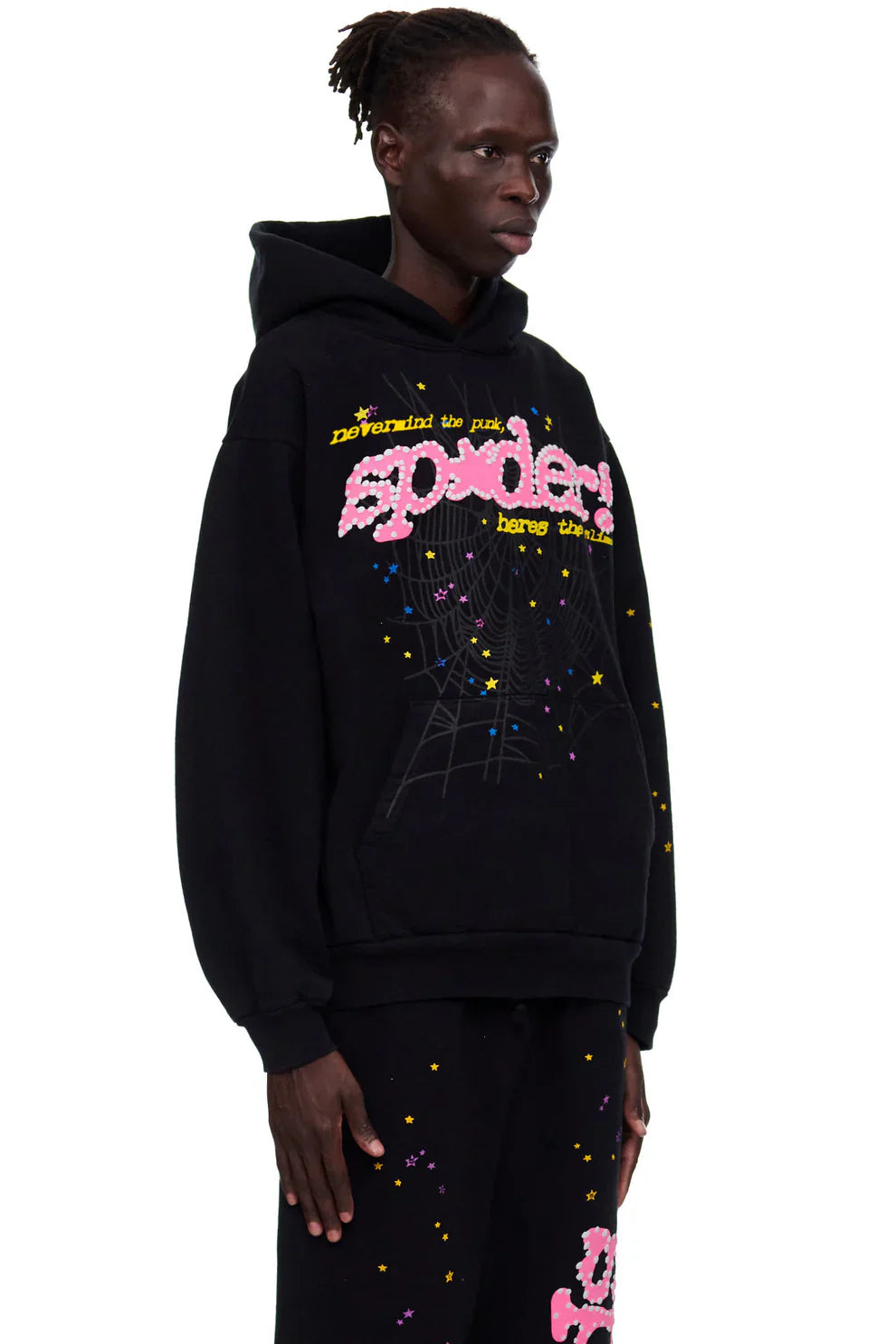 Wearing the Sp5der “Hares the Slime” Hoodie—a black 440GSM heavyweight streetwear pullover with star and web graphics—paired with matching black pants, the person stands against a plain white background.