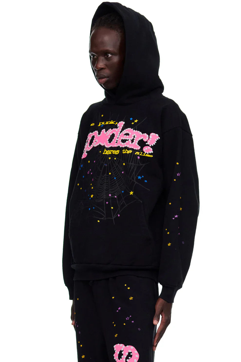 A person in the Sp5der “Hares the Slime” Hoodie – Black 440GSM Heavyweight Spider-Web Streetwear Pullover, featuring colorful star and spiderweb designs plus "a pink spider!" text, stands sideways.