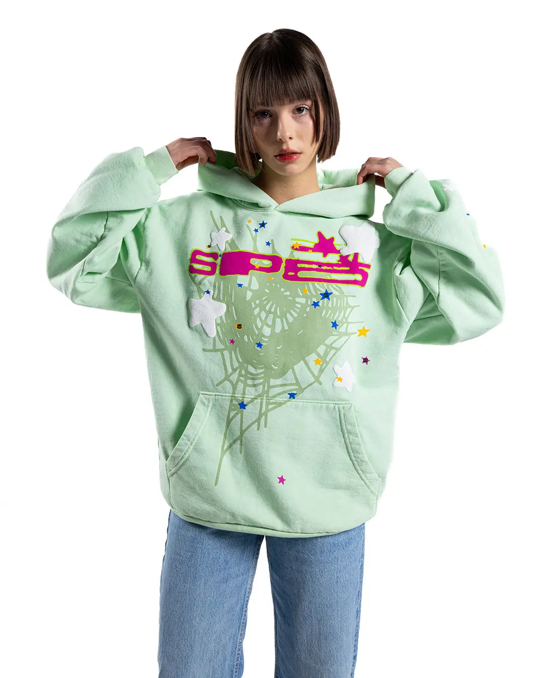 A person in a pastel green Sp5der “Mint Star Web” Hoodie – Mint Green 440GSM Heavyweight Oversized Streetwear Pullover with colorful designs, holding the hood, stands against a white background wearing light blue jeans.