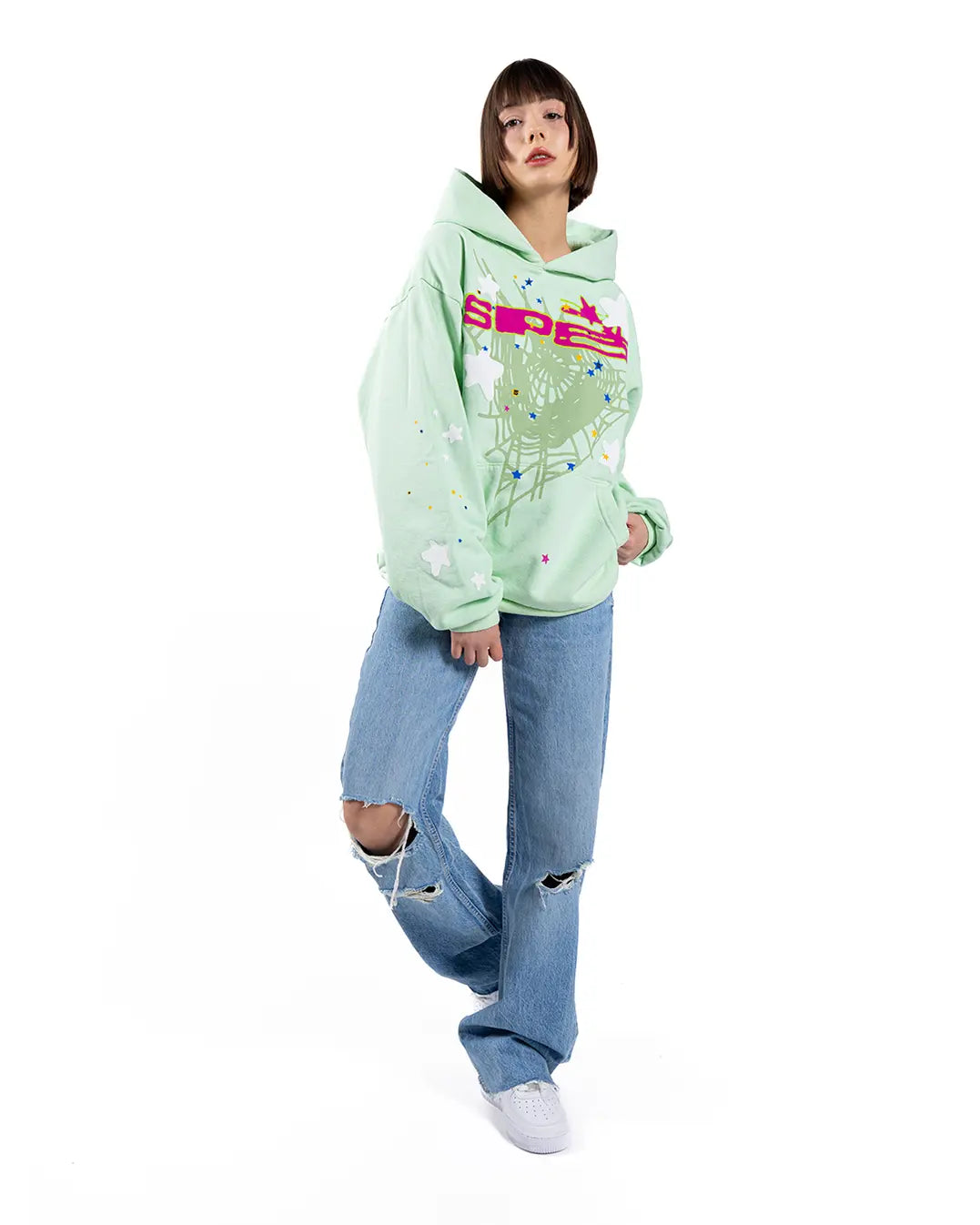 Someone wearing a Sp5der “Mint Star Web” Hoodie – Mint Green 440GSM Heavyweight Oversized Streetwear Pullover pairs it with blue ripped jeans and white sneakers, standing against a white background.