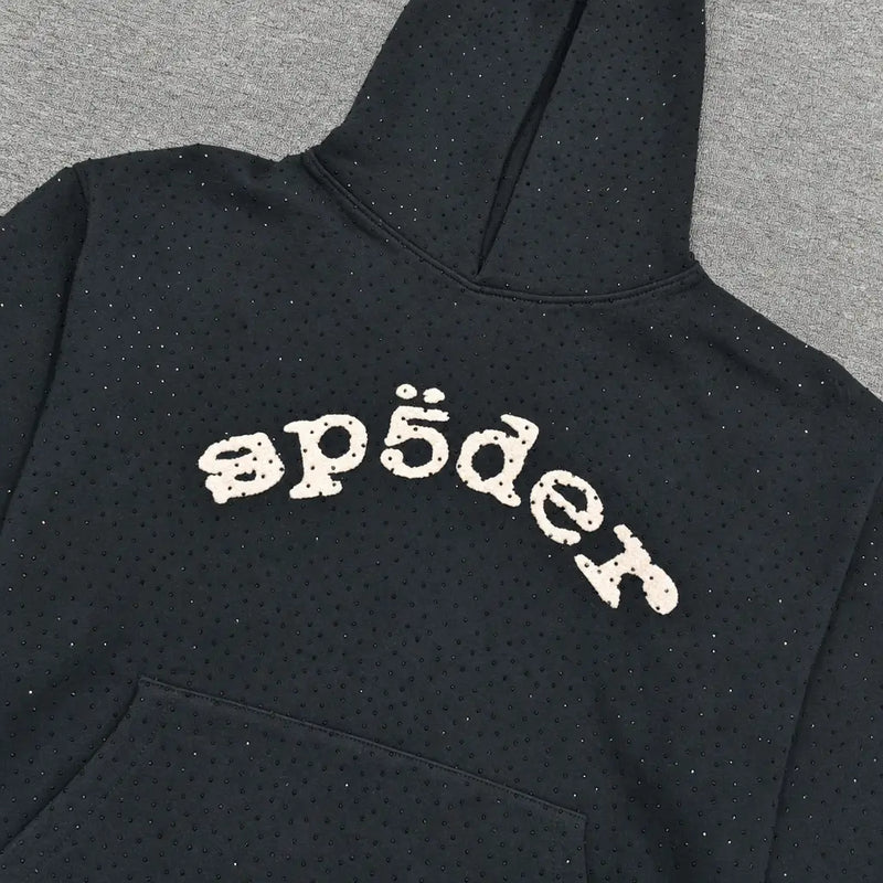 The Sp5der Black Rhinestone Hoodie Cream Arch Logo Cotton features subtle rhinestone embellishments and a stylized “sp5der” arch logo on the chest, laid flat on a gray surface to showcase its streetwear style.