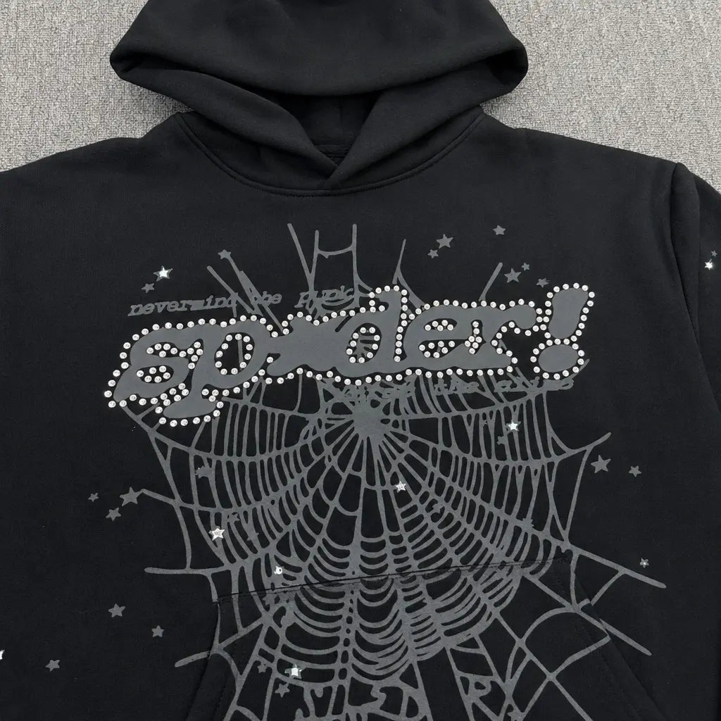 The Sp5der Black Rhinestone Spiderweb Graphic Hoodie showcases a silver spiderweb with sparkling accents, rhinestone logo, bold dotted "spider!" text on the chest, and floating stars and "never in the hell" above—ideal for streetwear style.