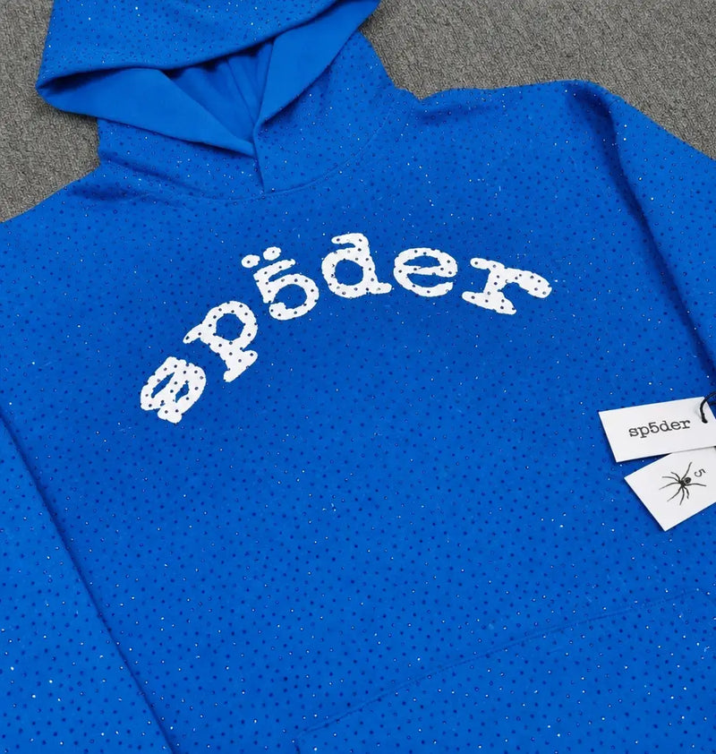 The Sp5der Blue Rhinestone All-Over Arch Logo Hoodie is a blue streetwear hoodie with silver rhinestones and “spõder” in white, stylized text across the chest, plus a tag featuring the same text and a spider illustration.